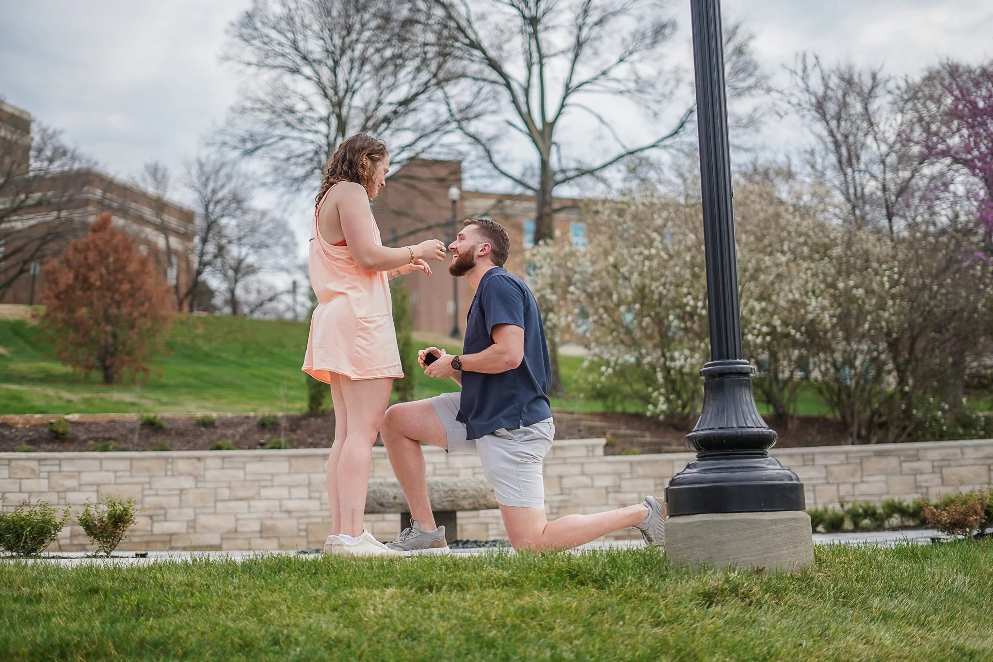 A man kneeling on one knee in front of a woman, proposing marriage in a park with trees and a building in the background.