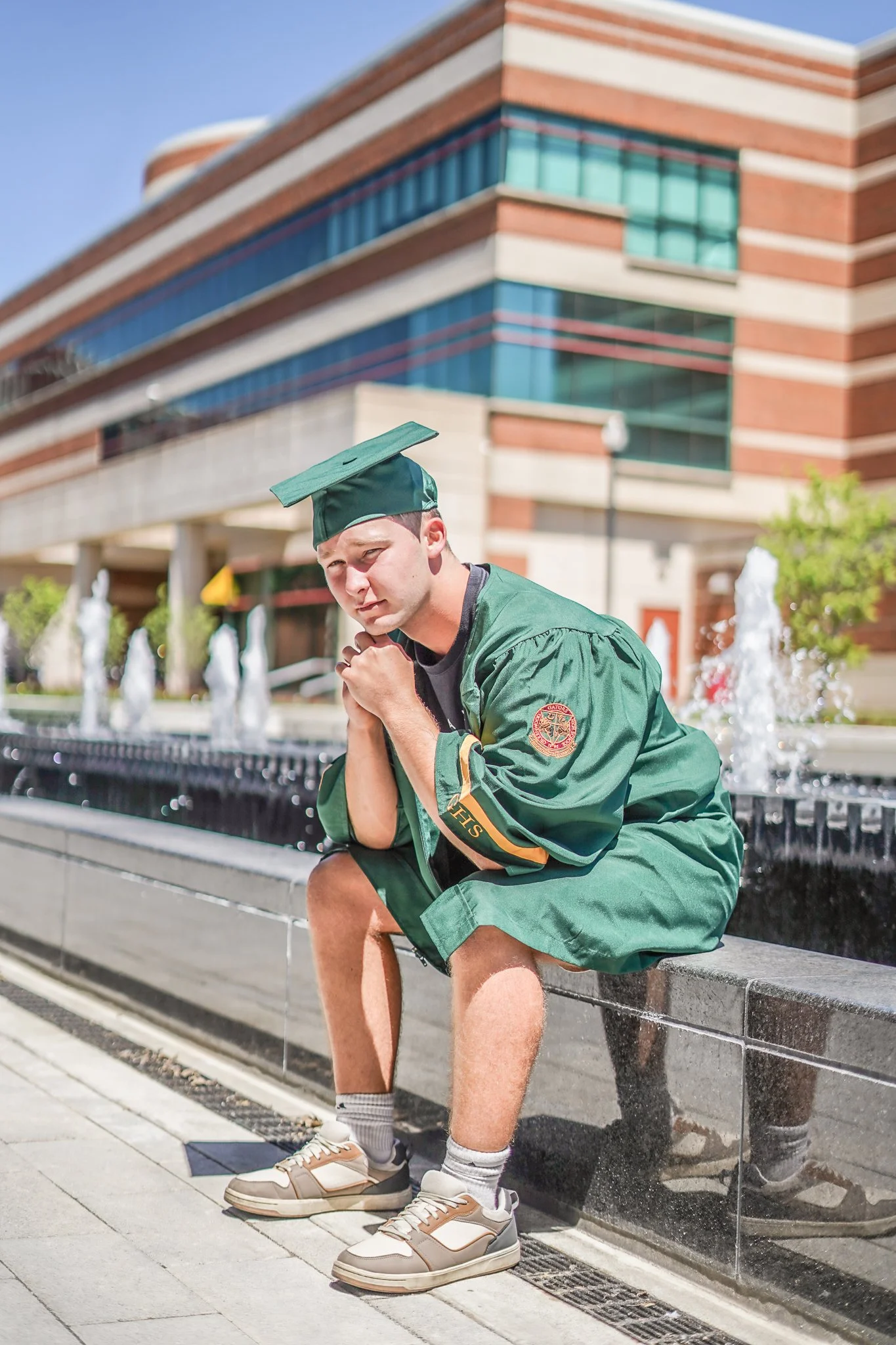 A young man in a green graduation cap and gown sitting on a bench outside a modern brick and glass building with fountains in the background, during daytime.