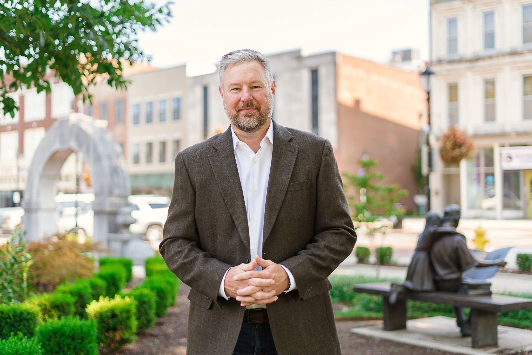 A middle-aged man with gray hair and a beard, wearing a dark gray blazer and white shirt, standing outdoors in a city park with trees, benches, and buildings in the background.