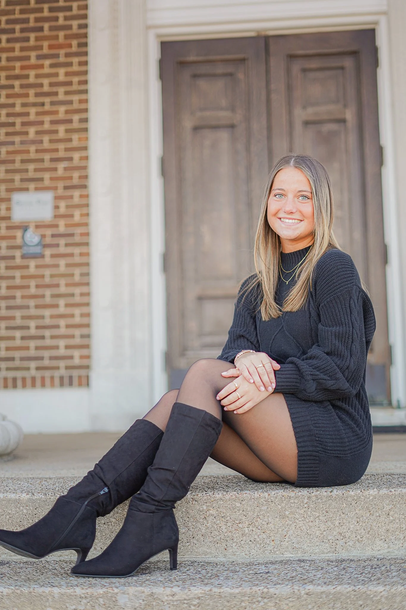 A woman with blonde hair, blue eyes, wearing a black sweater, black thigh-high boots, and jewelry, sitting on concrete steps in front of a brick building with a wooden door, smiling at the camera.