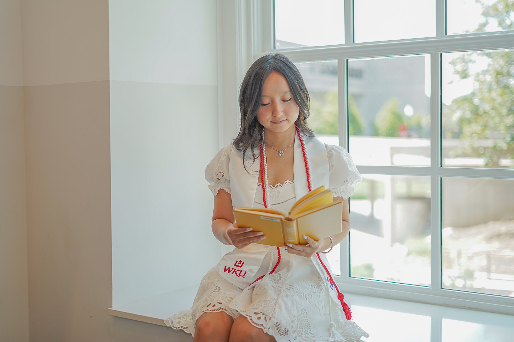 A woman wearing a white dress with lace and a red sash reading 'WKU' sitting on a windowsill, reading a yellow book.