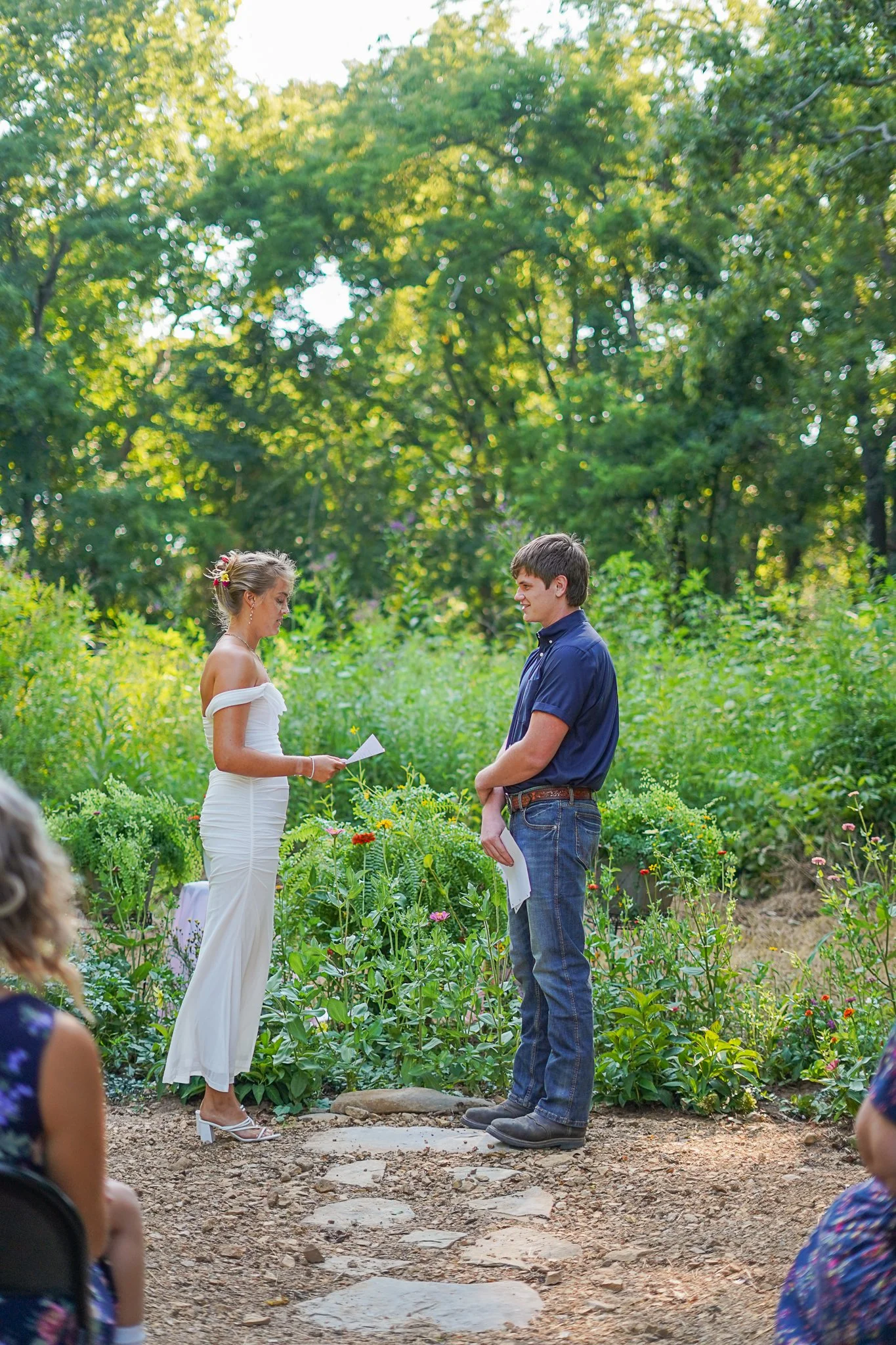 A couple stands outdoors in front of a green garden, exchanging vows during a wedding ceremony on a sunny day.