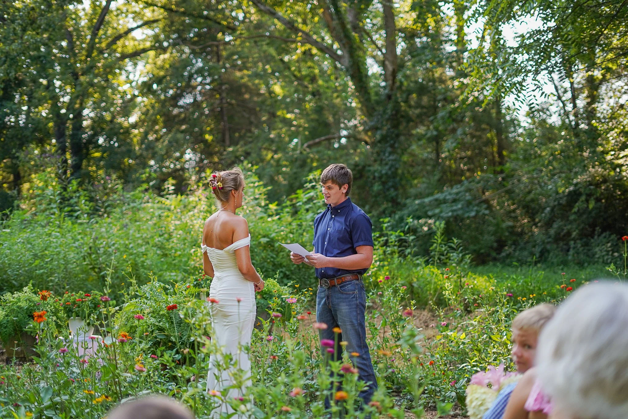 A couple getting married in a garden, with the officiant reading from a paper, surrounded by flowers and trees.