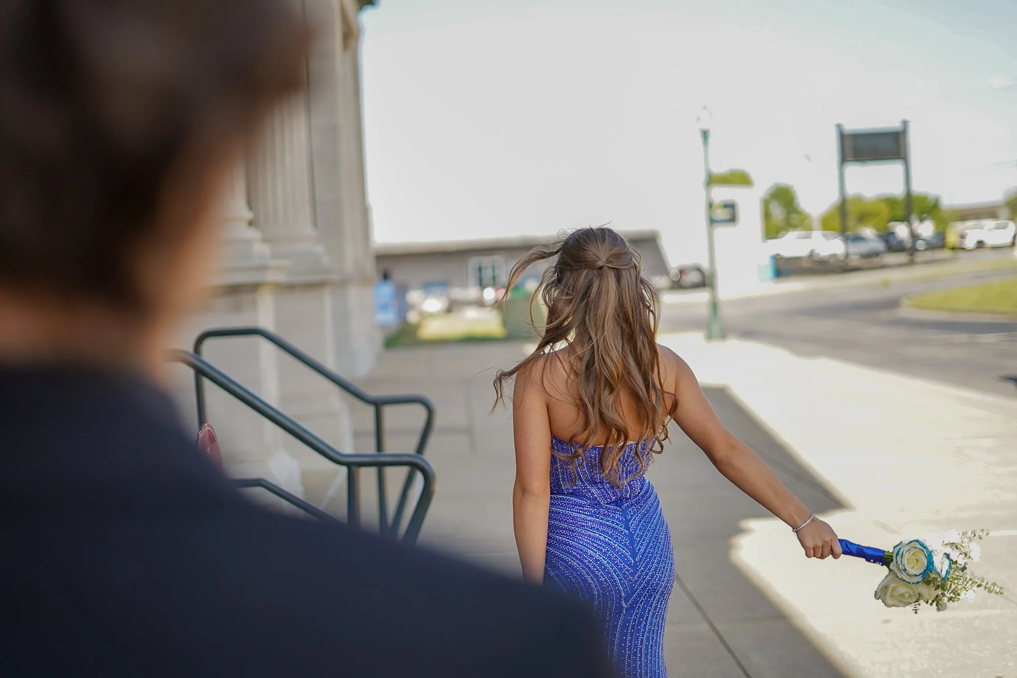 A woman in a blue dress holding a bouquet of white roses is walking on the sidewalk outside a building as the sun shines brightly.