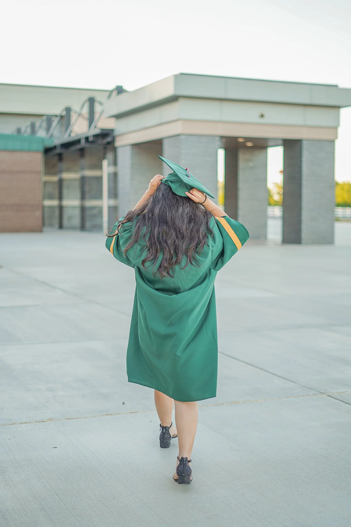 A person dressed in a green graduation gown and cap, walking away on a wide concrete area outside a modern building, holding their cap with both hands.