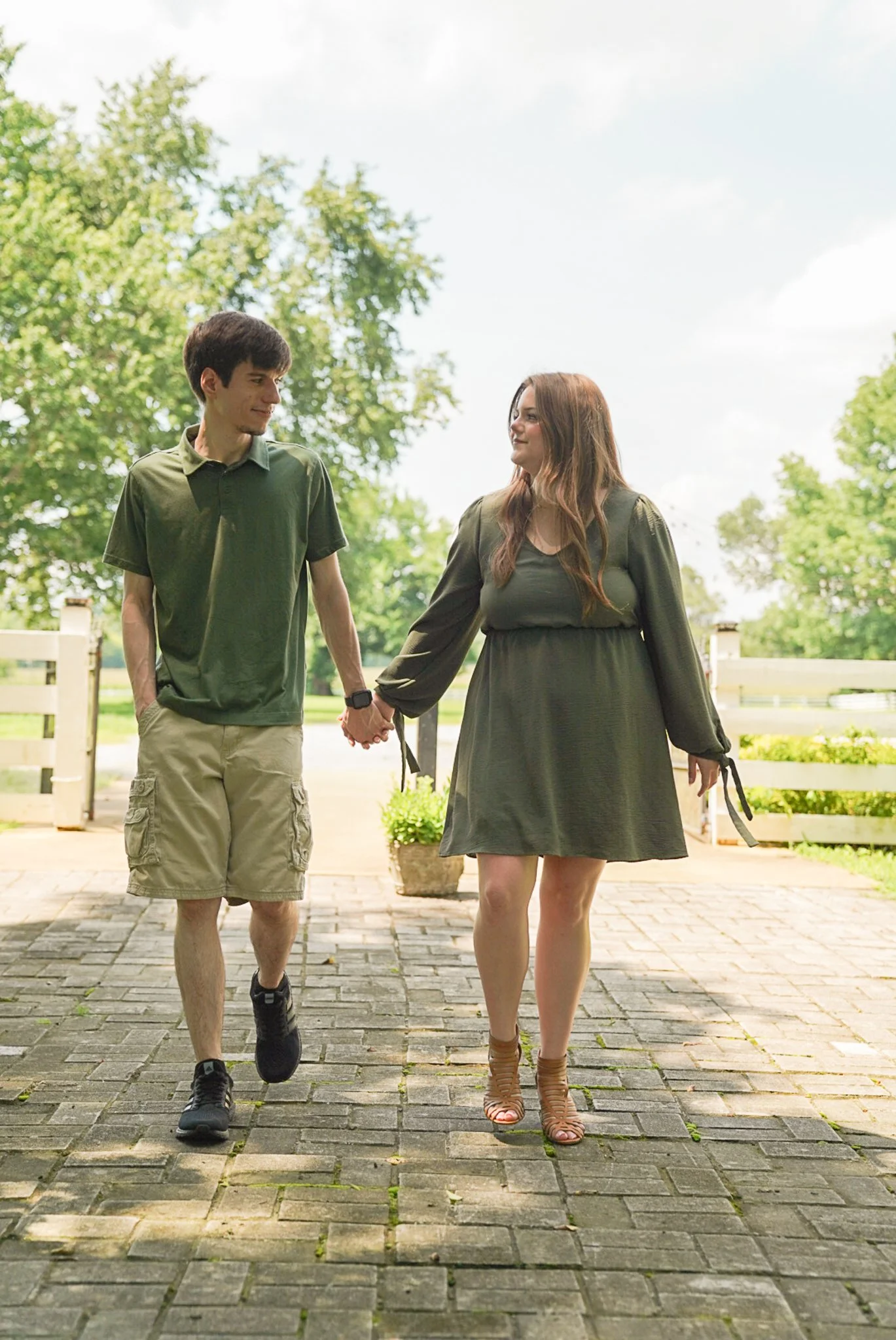 A young man and woman walk hand-in-hand on a paved path outdoors, surrounded by green trees and a white fence, enjoying a sunny day.