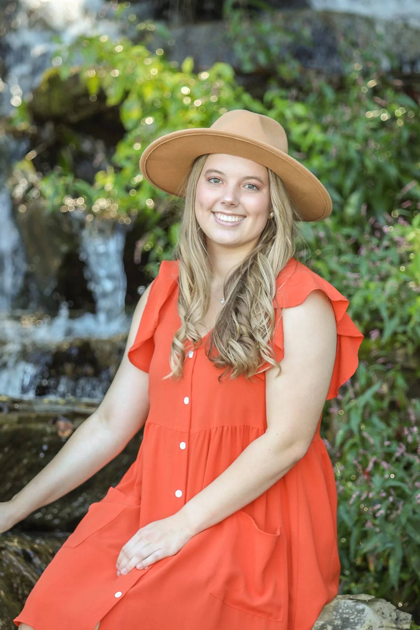 Young woman with long blonde hair wearing a wide-brimmed tan hat and an orange sleeveless dress with white buttons, sitting outdoors near a small waterfall with lush green foliage in the background.