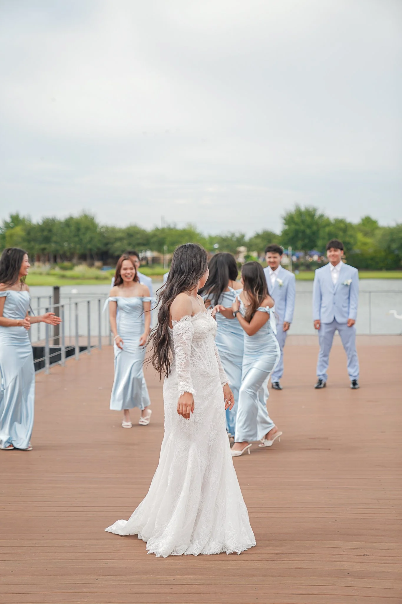 A bride in a white lace wedding dress stands on a wooden deck, with bridesmaids and groomsmen in matching light blue dresses and suits in the background near a body of water, celebrating outdoors on a cloudy day.
