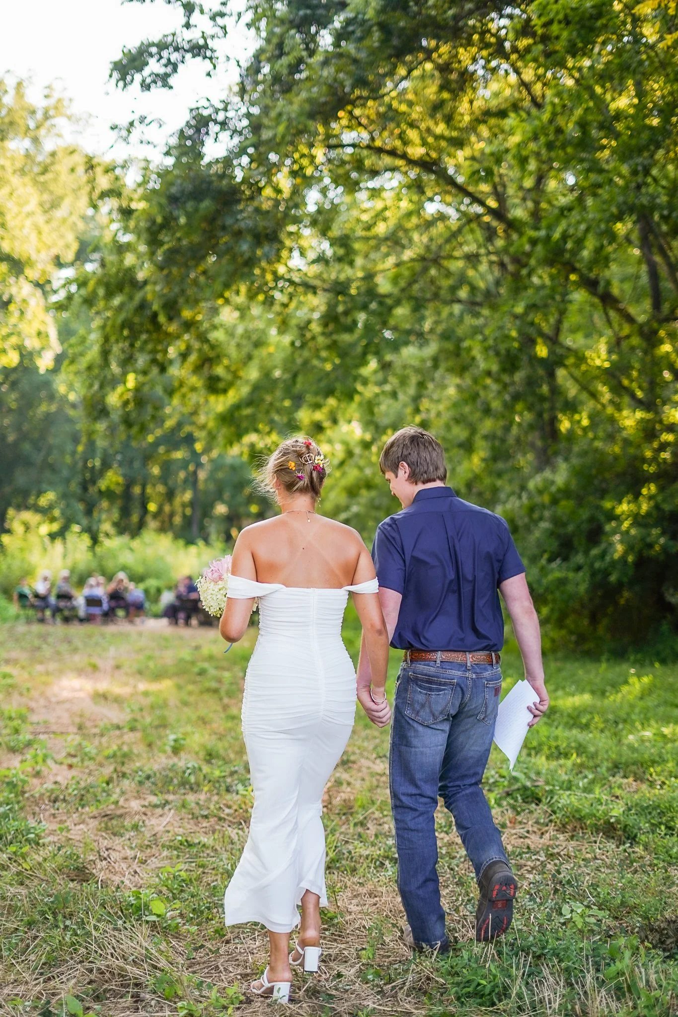 A newlywed couple walks hand-in-hand in a lush green park, with guests seated on benches in the background, celebrating their wedding.