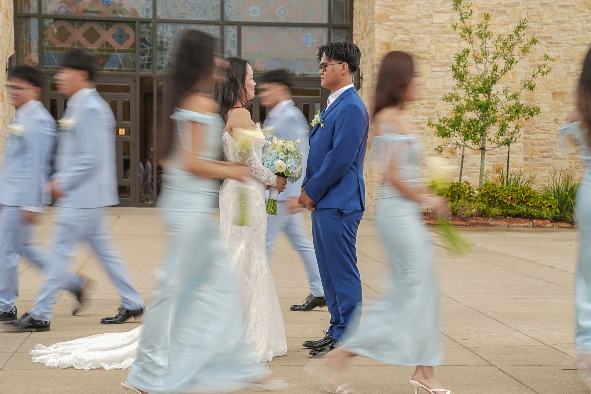 A bride and groom standing face to face during their wedding ceremony with blurred bridesmaids and groomsmen walking in front of them.