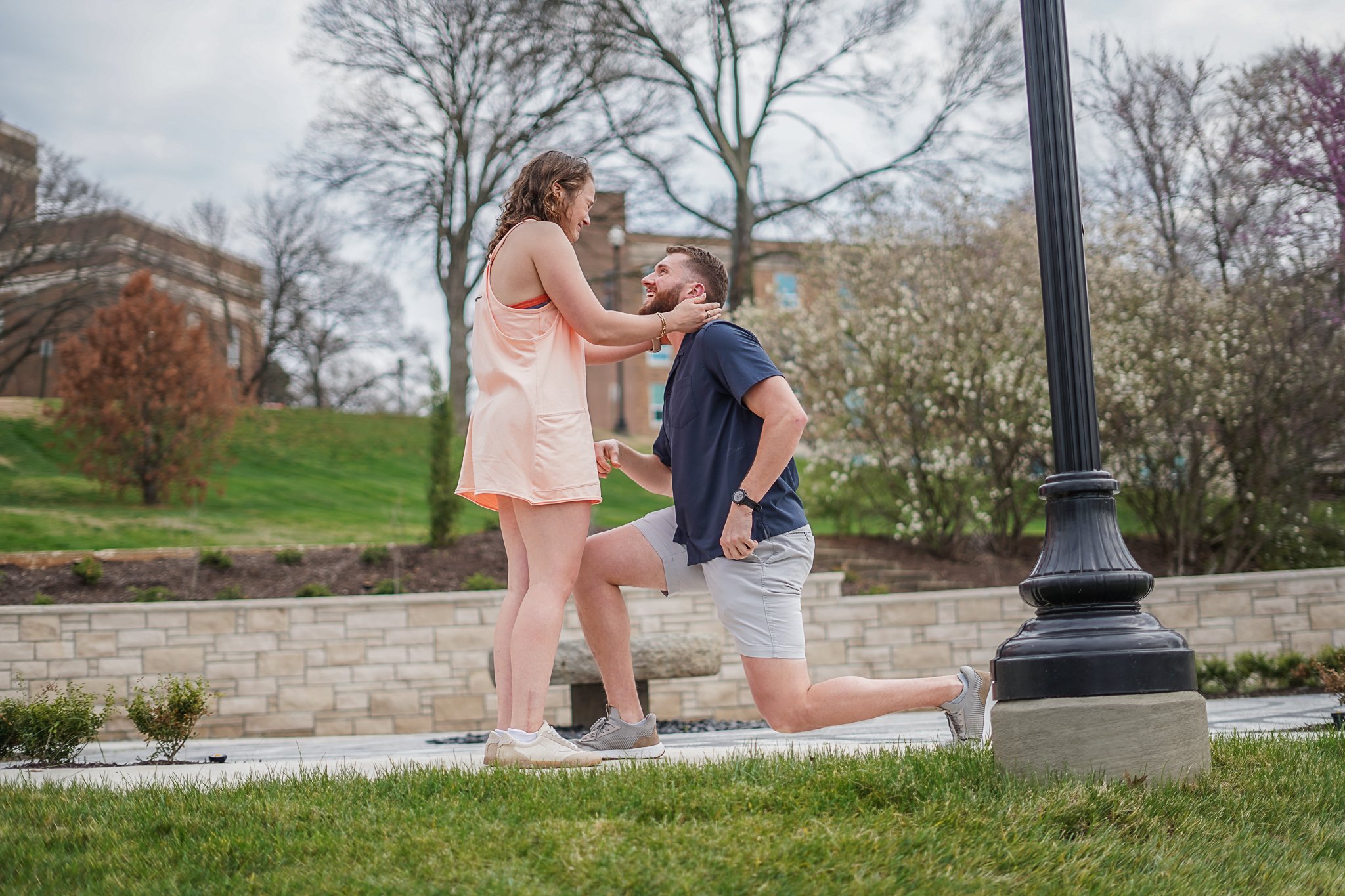 A woman in a peach tank top and shorts is holding a man's face as he kneels with one knee on a step in a park, smiling at her. The man is wearing a navy shirt and shorts. They are near a large black lamp post with trees and a park building in the bac