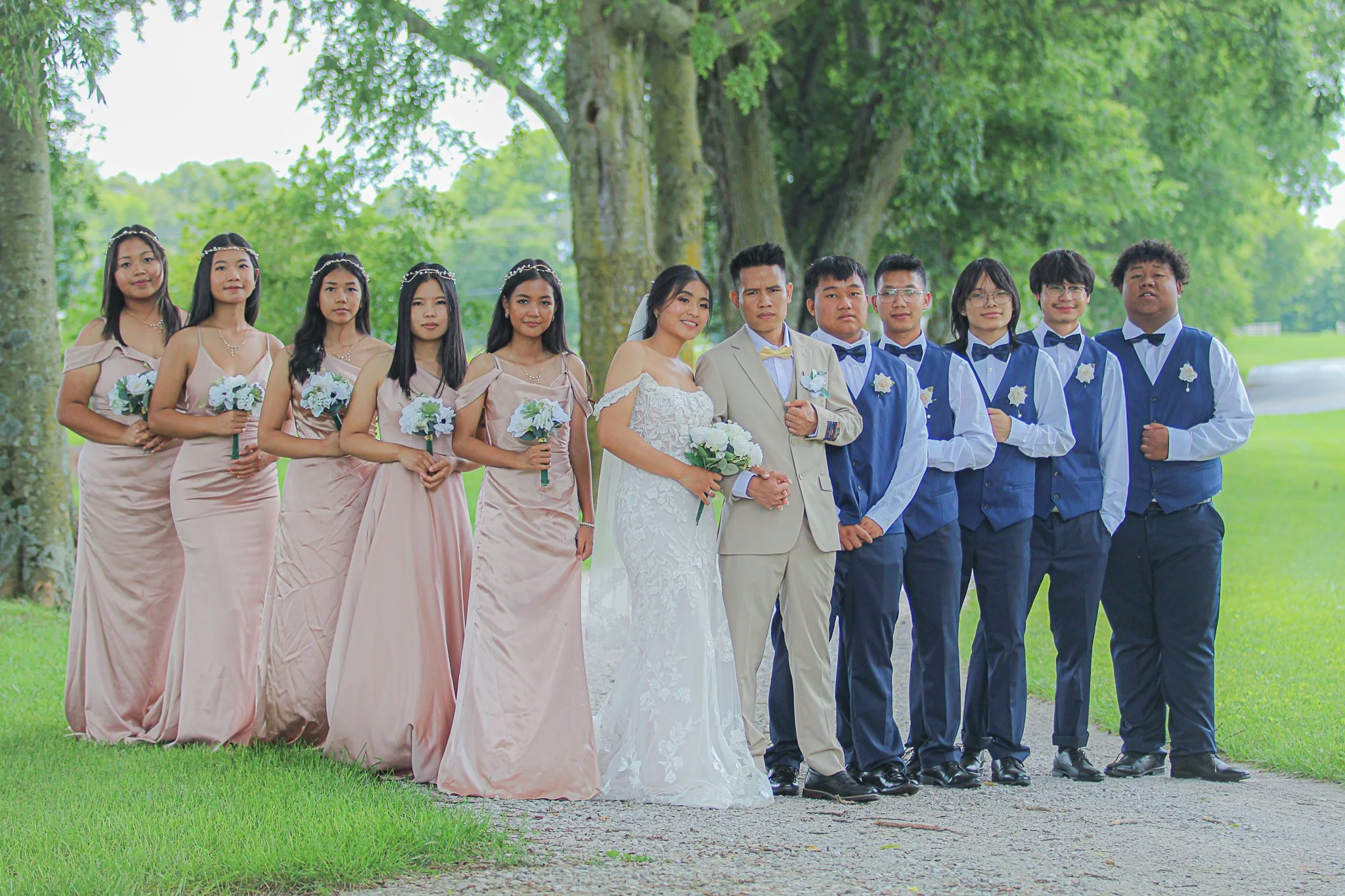 A group of eleven people dressed in formal wedding attire, standing in a park with green trees and grass.