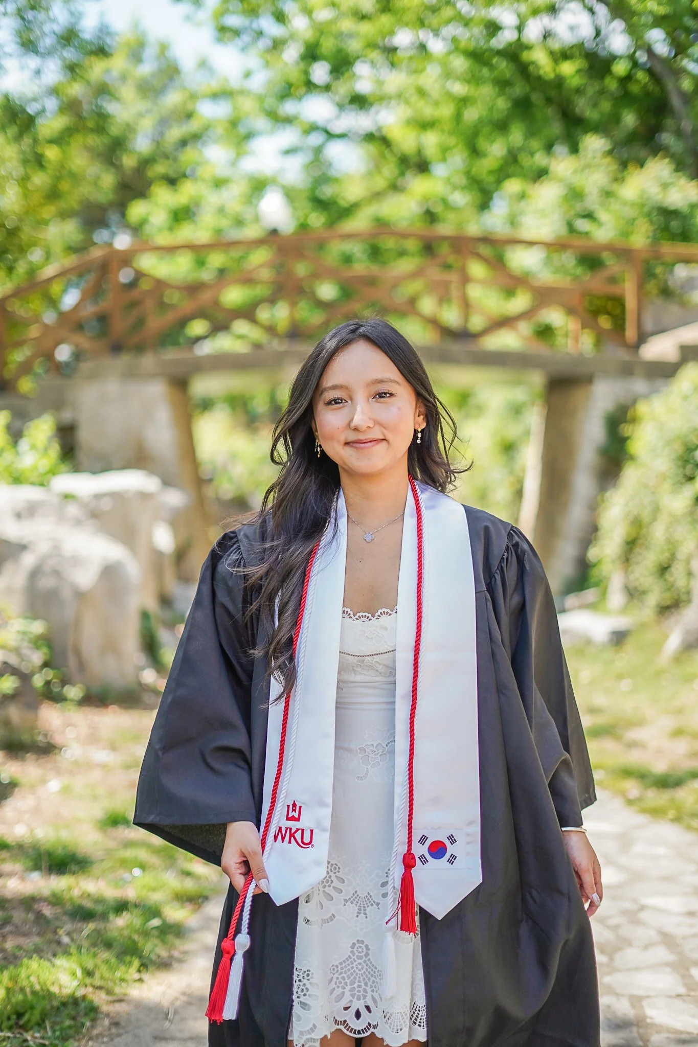 A young woman wearing a graduation cap and gown, a white stole with a red and white cord, standing outdoors with trees in the background during daytime.