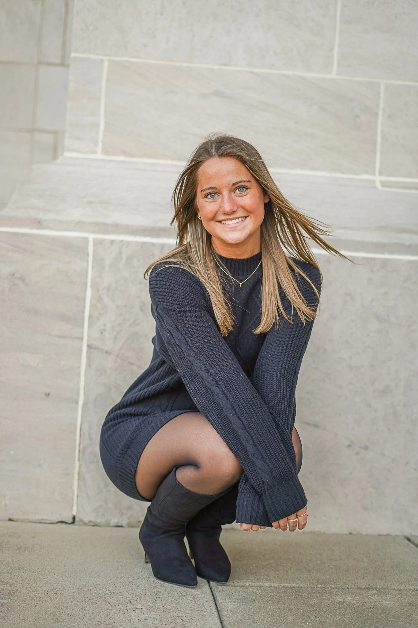 A young woman with long, light brown hair, blue eyes, and a big smile poses against a stone wall. She is wearing a black long-sleeve top, a necklace, rings, and black pants, with her knees bent and arms crossed over her knees.