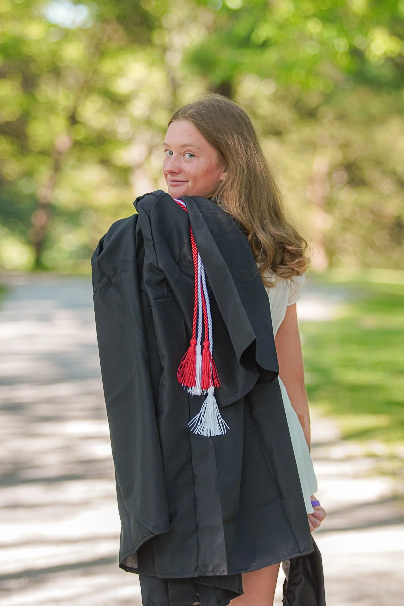 A young woman with brown hair, wearing a graduation gown and sash, standing outdoors with a blurred green background, smiling at the camera.