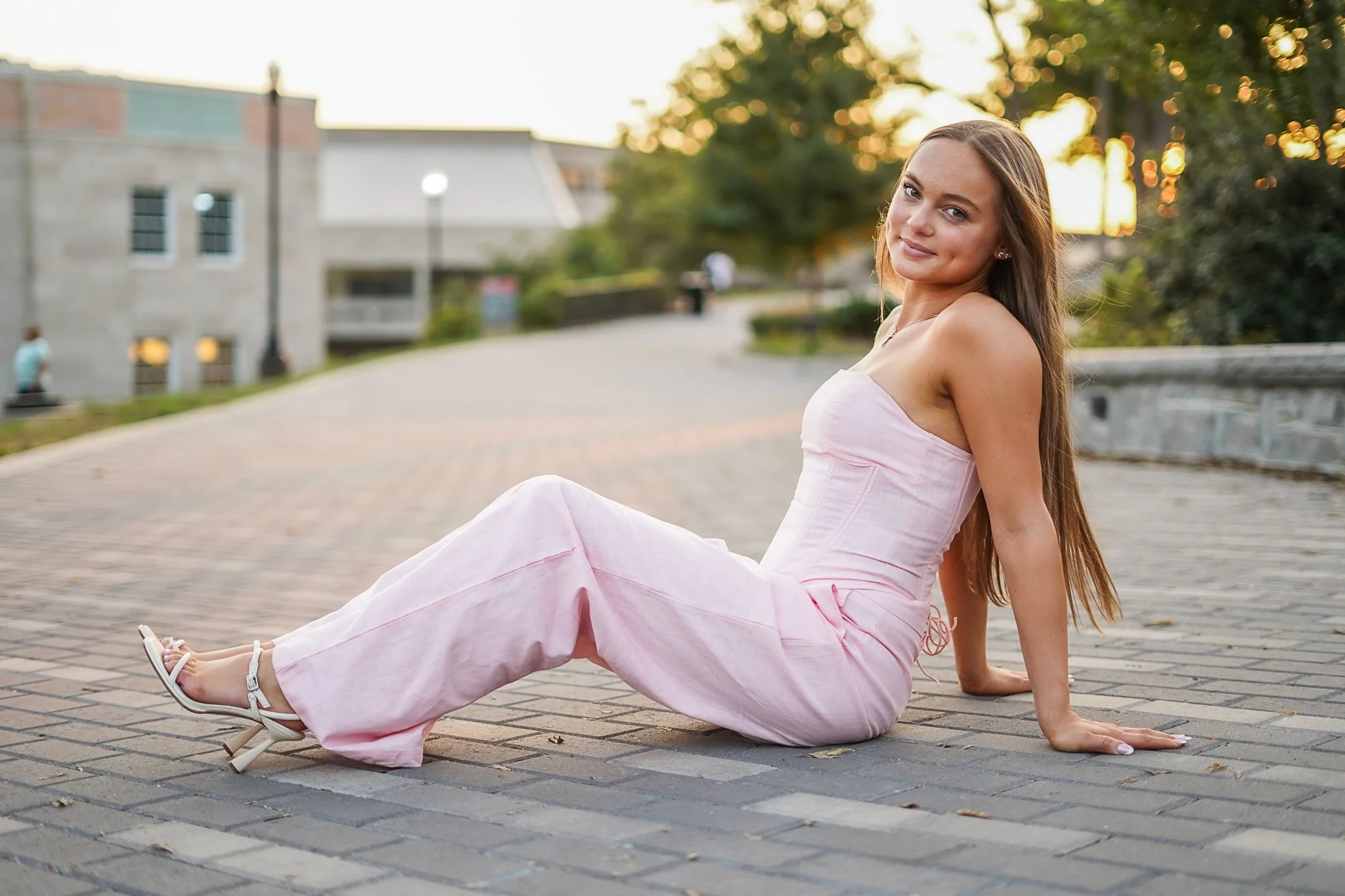 A young woman in a pink dress sitting on a brick sidewalk during sunset, smiling at the camera with her legs stretched out and her arms supporting her on the ground. Background includes a building, trees, and street lamps.