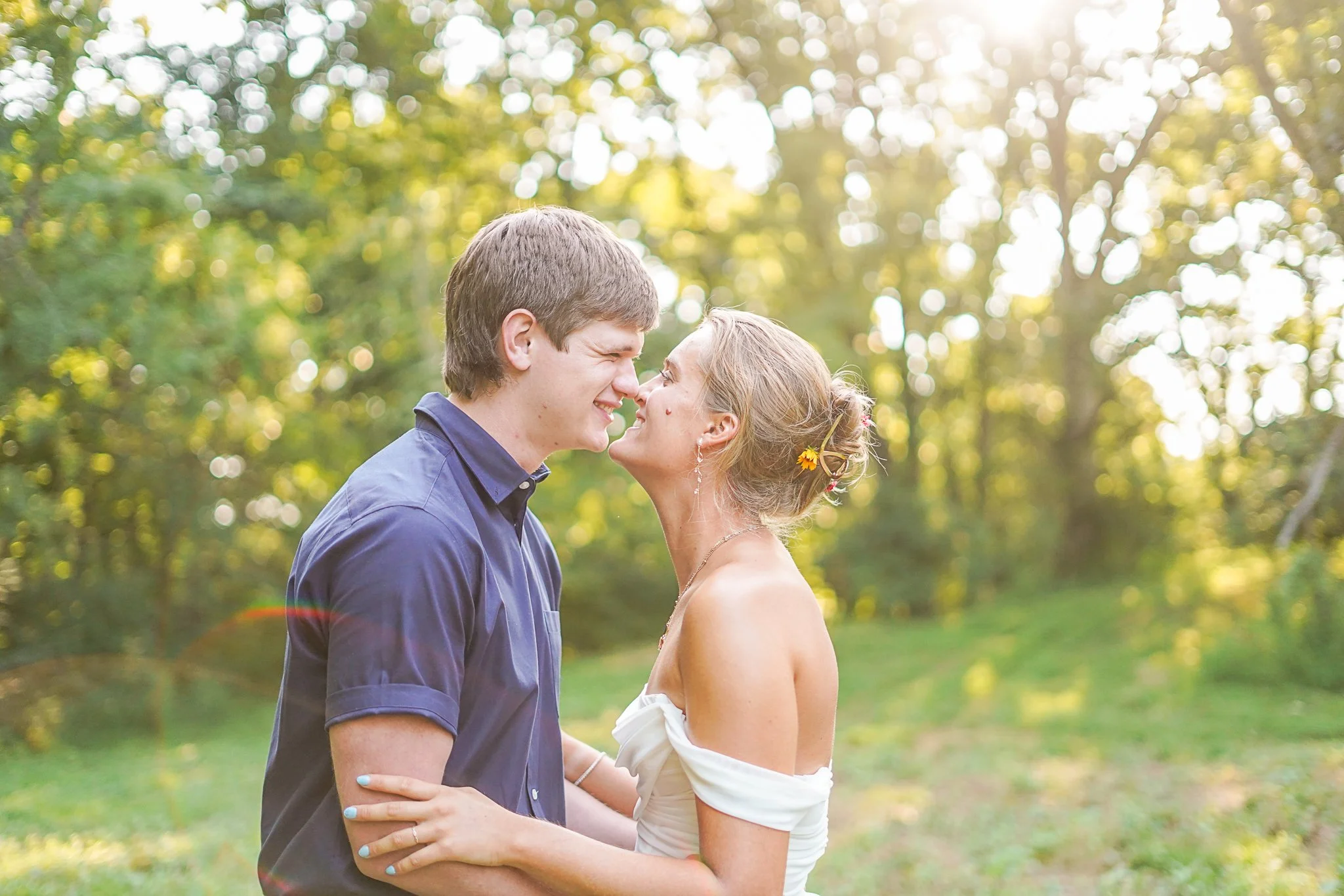 A young couple is standing close together outdoors in a park with sunlight filtering through green trees, smiling and about to kiss.