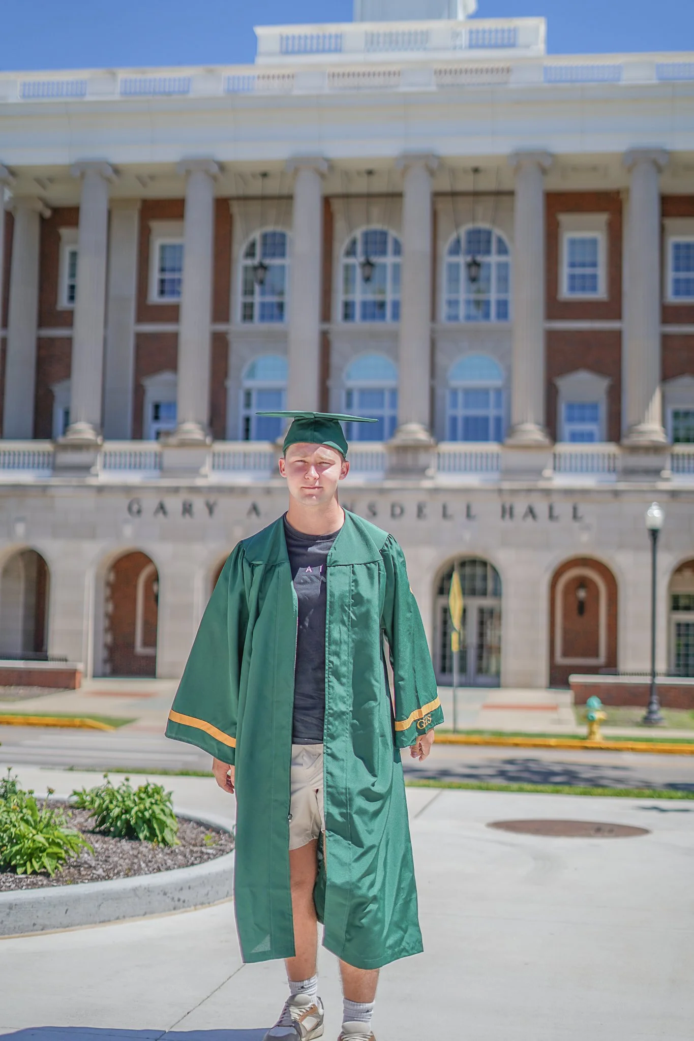 A young man in a green graduation gown and cap standing in front of a large historic building with columns and arched windows.