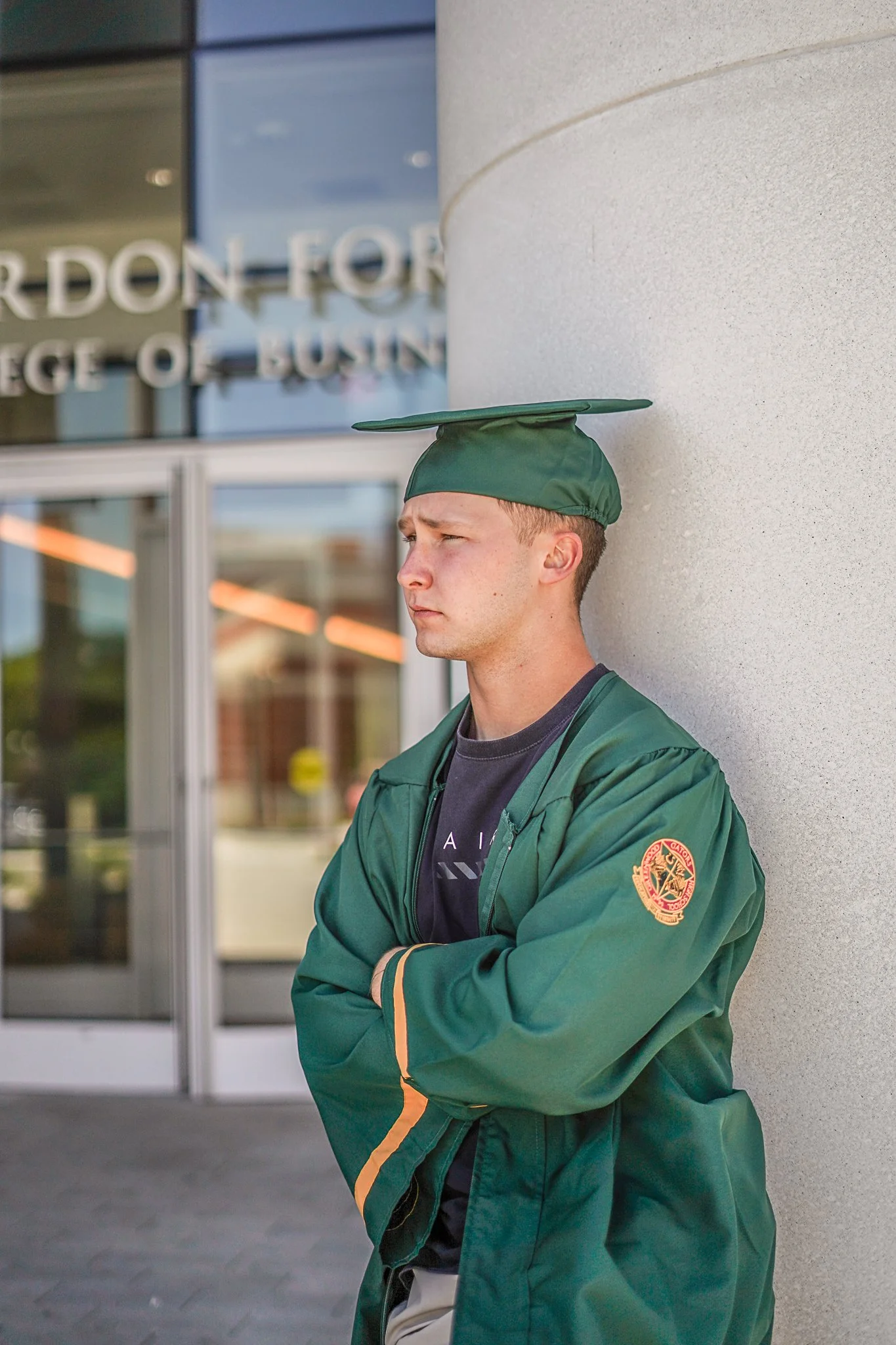 A young man with short hair and light skin wearing a green graduation cap and gown with a patch on the sleeve, standing outside a building with glass doors and a beige wall, looking to the side with arms crossed.