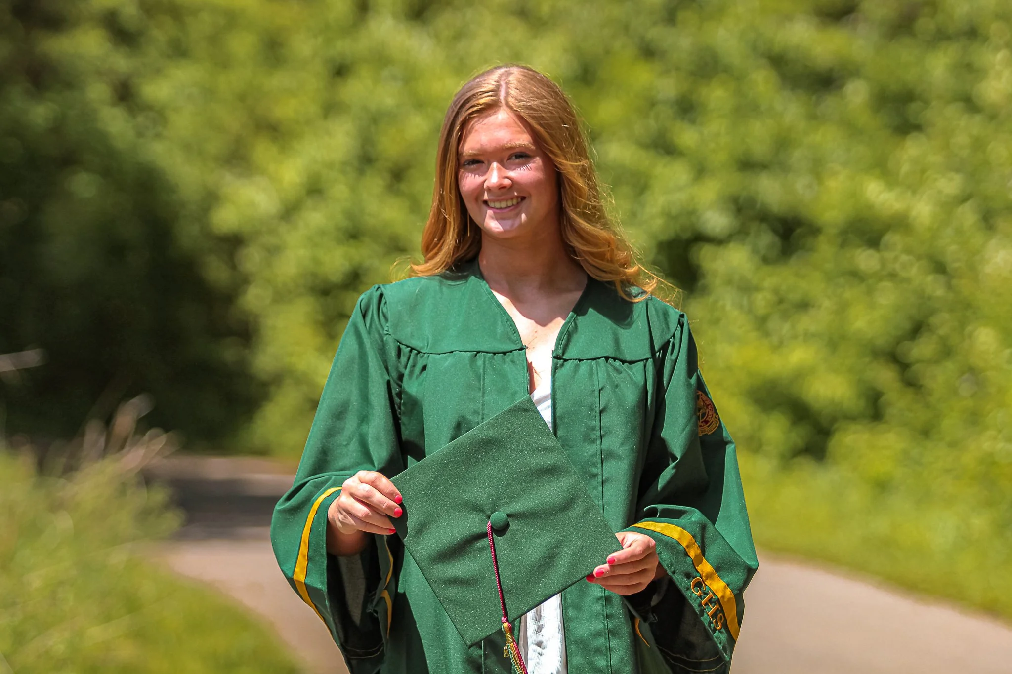 A woman in a green graduation gown holding her diploma outdoors with green foliage in the background.