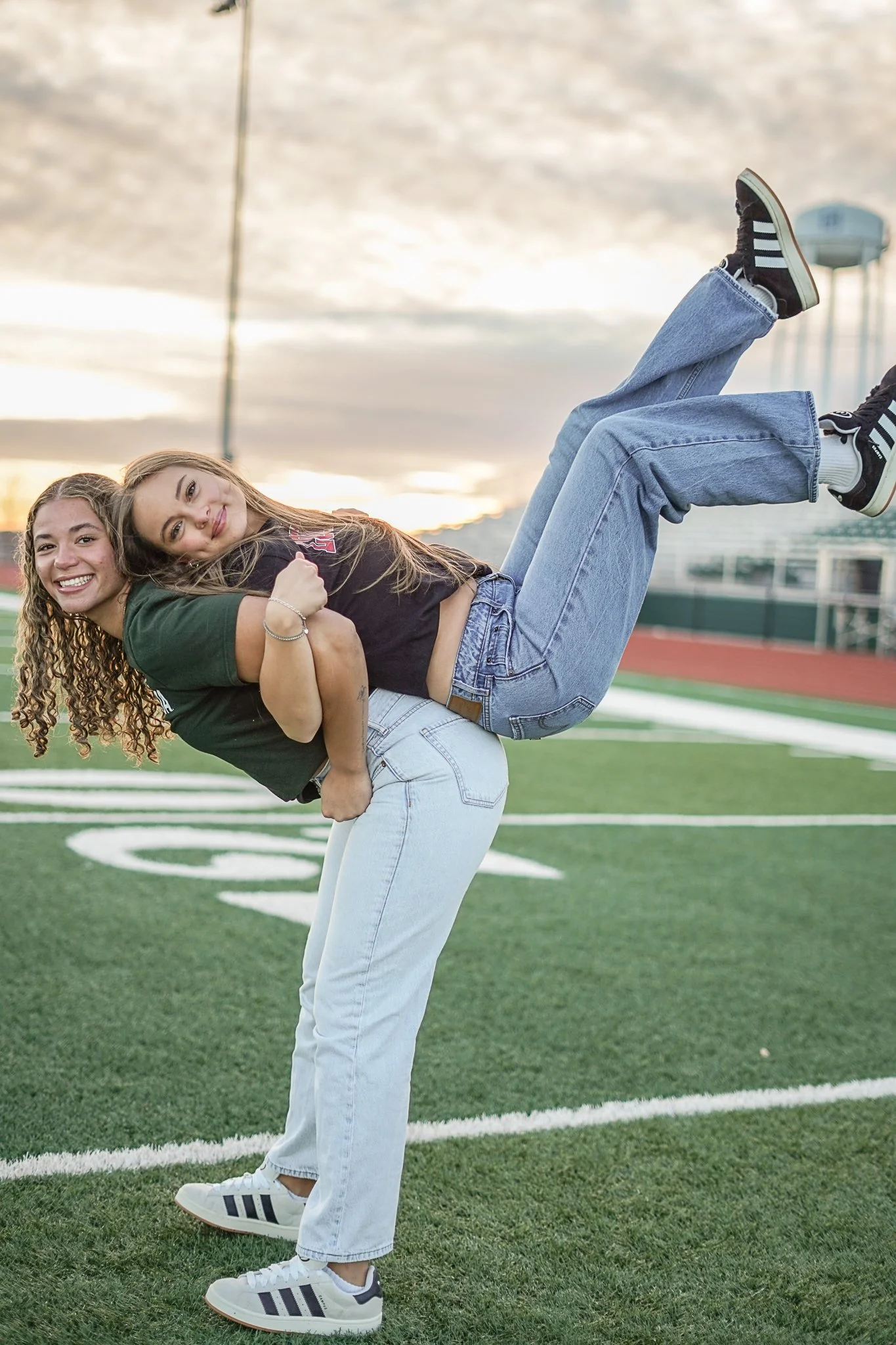 Two young women are standing on a football field, with one woman bent forward and the other woman lying back on her, as the woman in the back is holding her around the waist. The woman in the front has long, wavy blonde hair, is smiling, and is weari