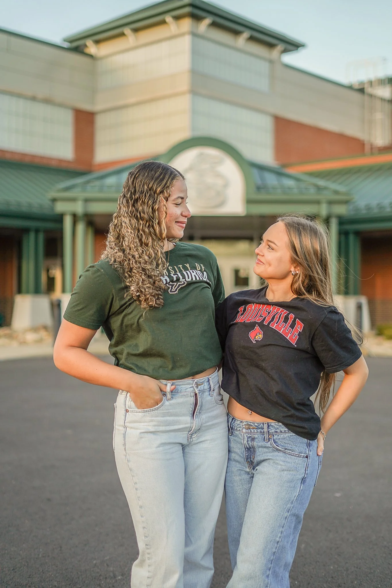 Two young women standing close together outside in front of a building, smiling and looking at each other. One woman has curly hair and is wearing a green T-shirt with 