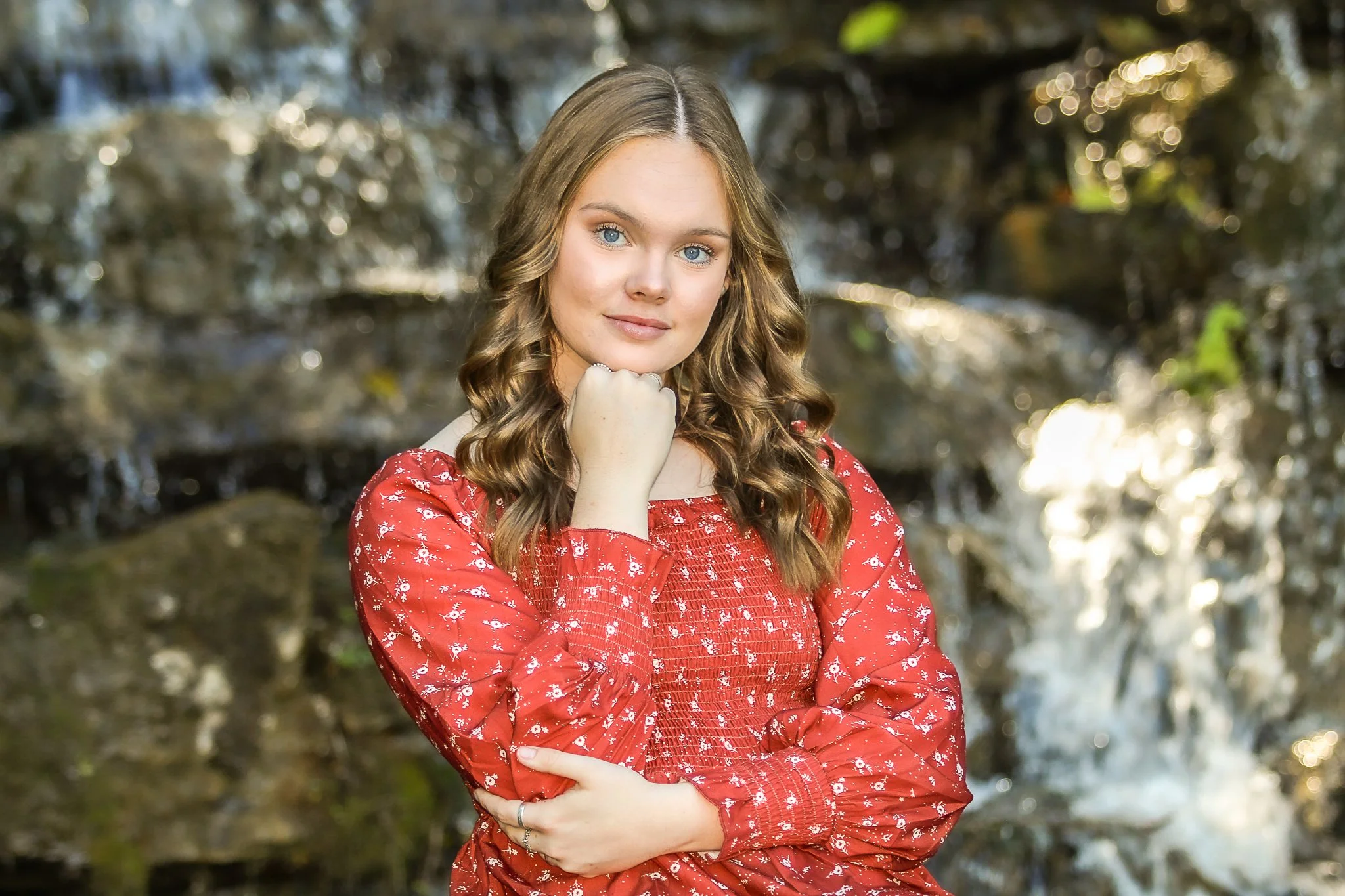 A woman with long, wavy blonde hair wearing a red dress with white floral pattern, posing outdoors with a natural background of blurred trees and water.