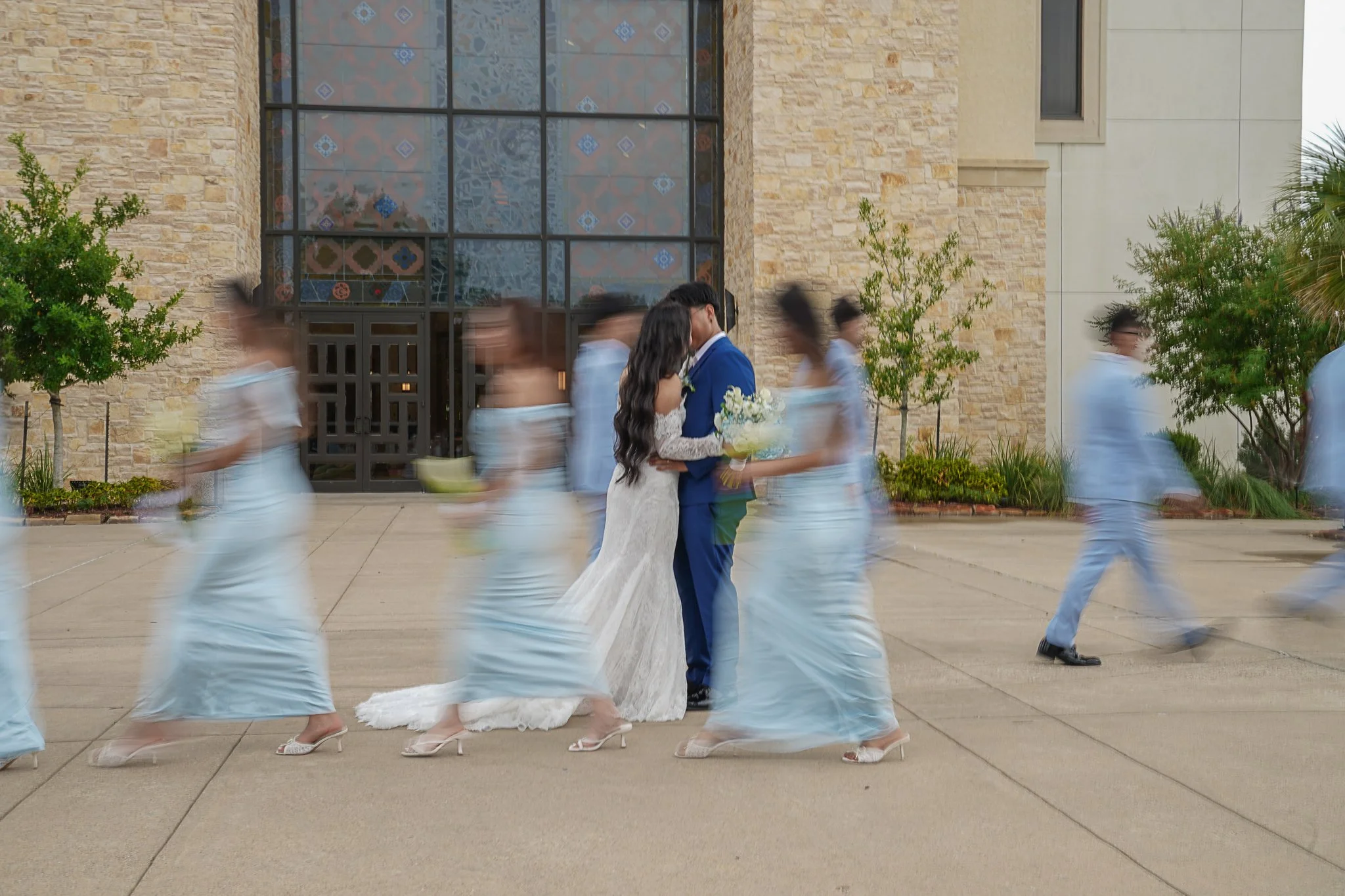 A bride and groom sharing a kiss while surrounded by women in blue dresses who are blurred from movement, at what appears to be a wedding. The scene is set outside a building with large windows and stone walls, with greenery on either side.