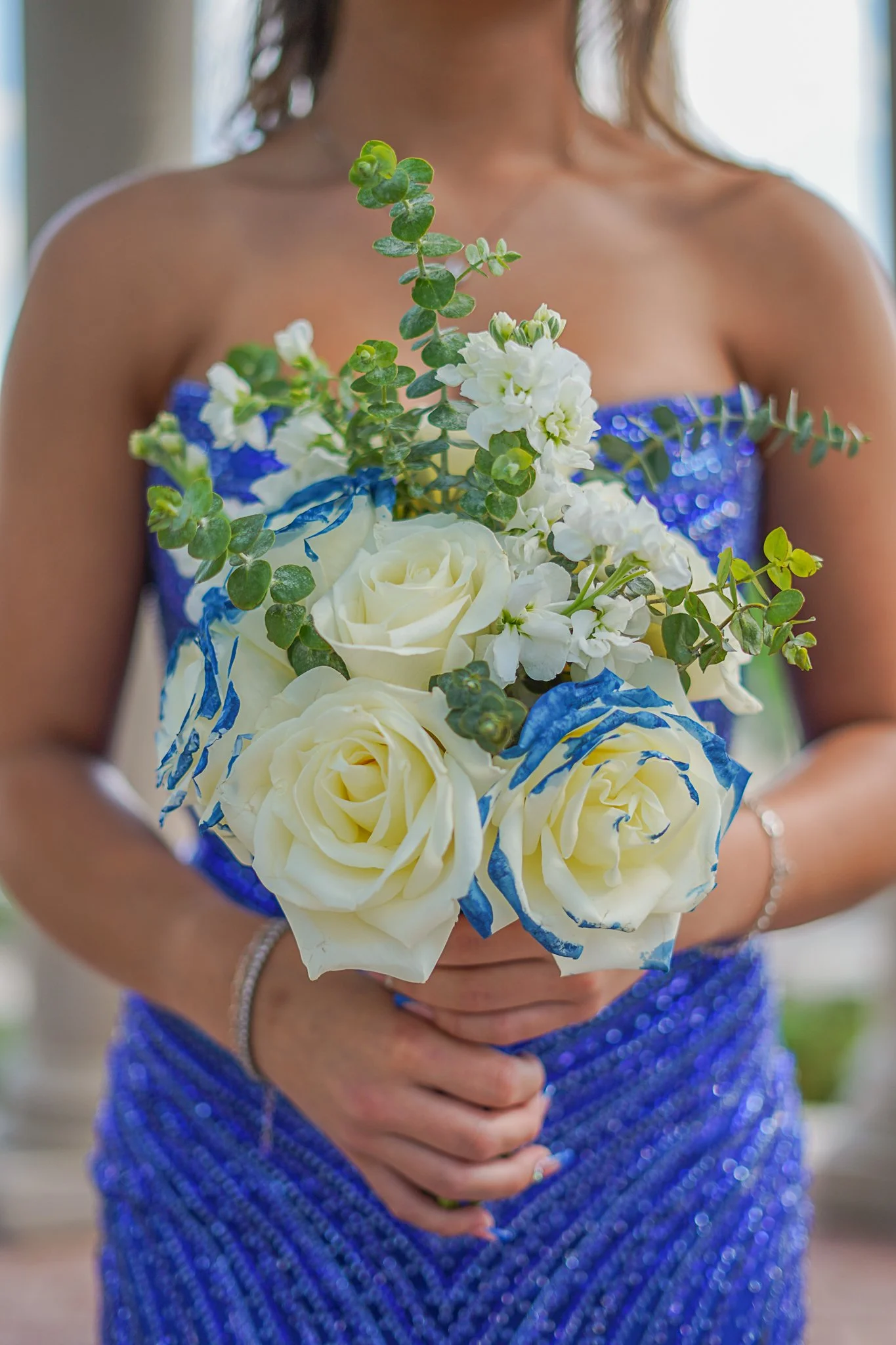 A person wearing a strapless blue dress with sequins holding a bouquet of white roses, green foliage, and blue ribbon.