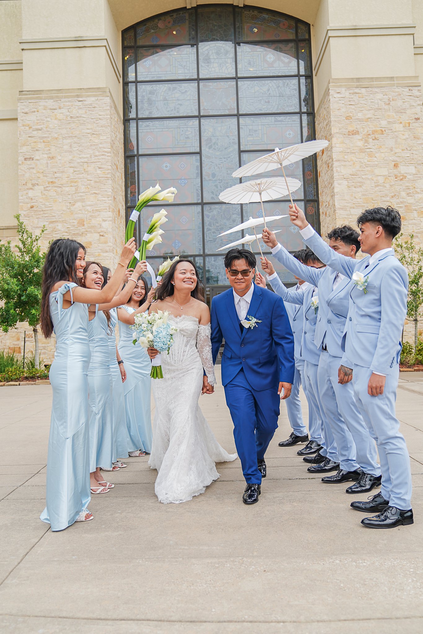 A bride and groom walk together while waiting outside a building with large stained glass window, surrounded by friends and wedding party holding white umbrellas and flowers.
