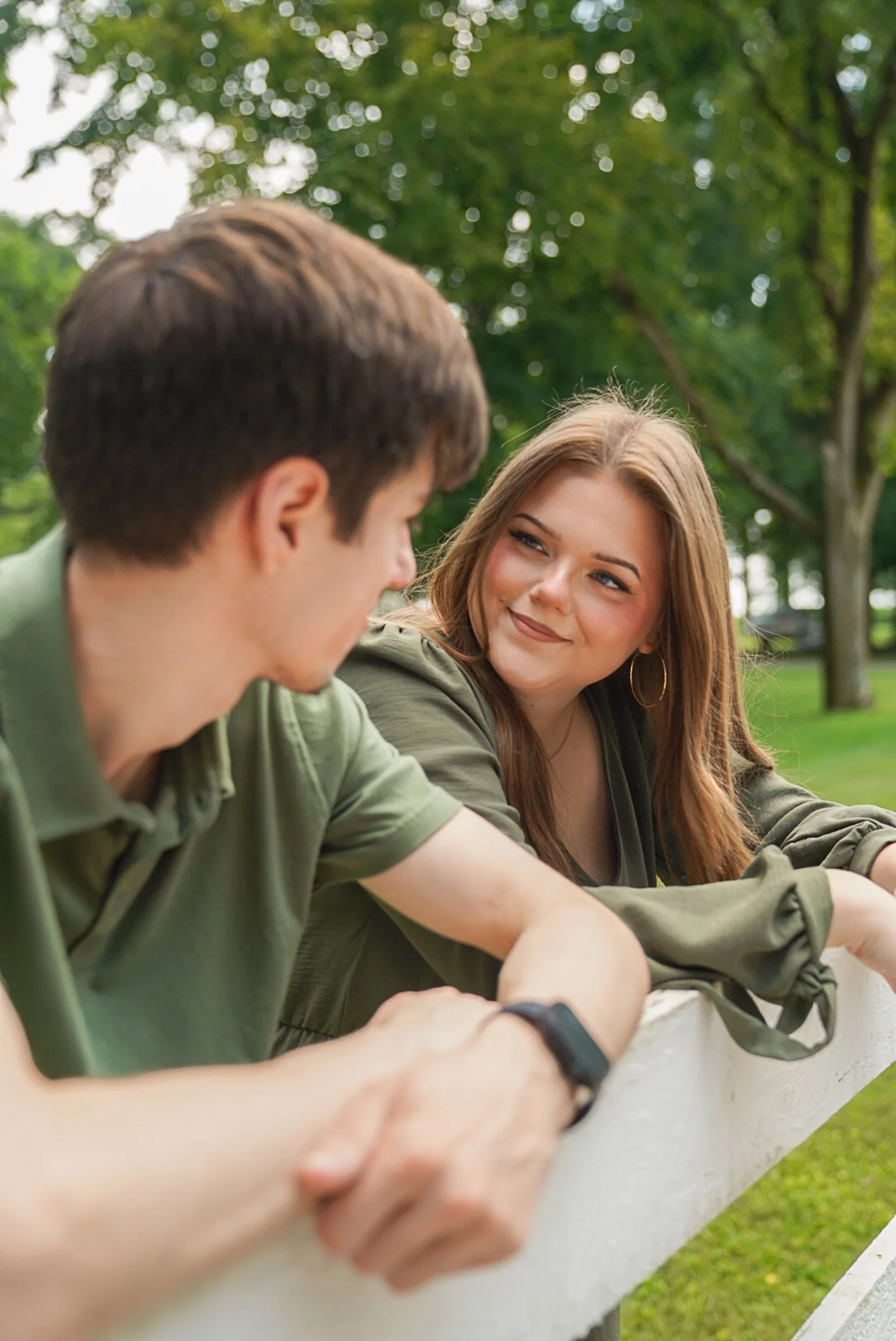 A young woman with long hair and earrings smiling at a young man with short hair. They are sitting outdoors at a park with green trees in the background, engaged in conversation.