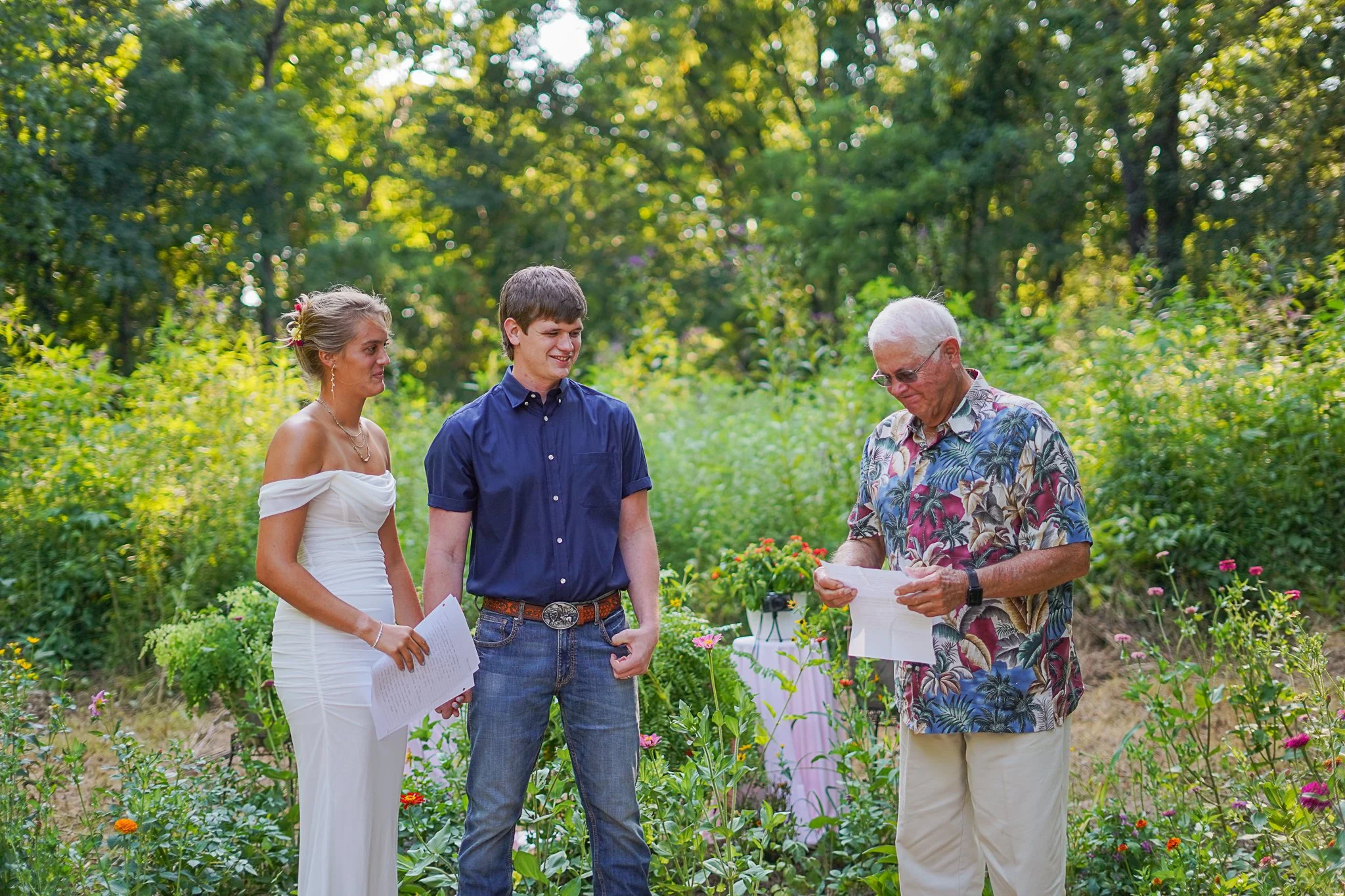 Three people, two men and a woman, standing in a garden with flowers, engaged in a conversation while holding papers, with trees and greenery in the background.