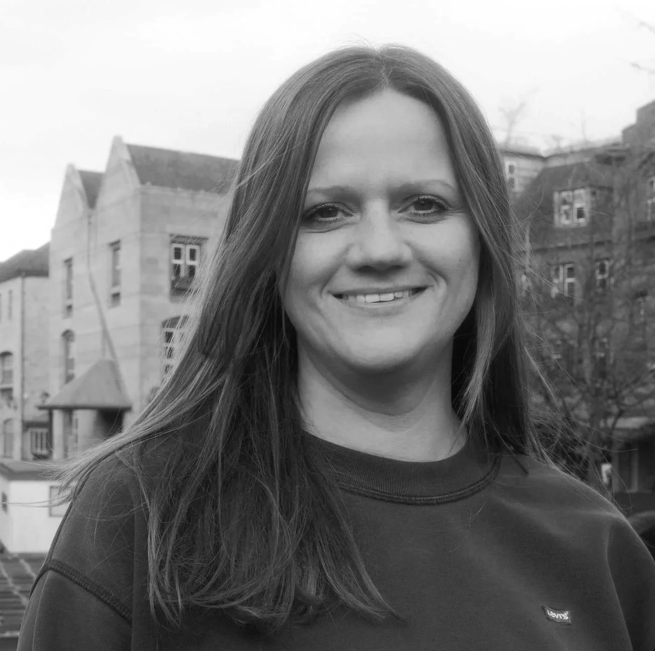 Black and white photo of a woman with long hair smiling outdoors, with buildings and trees in the background.