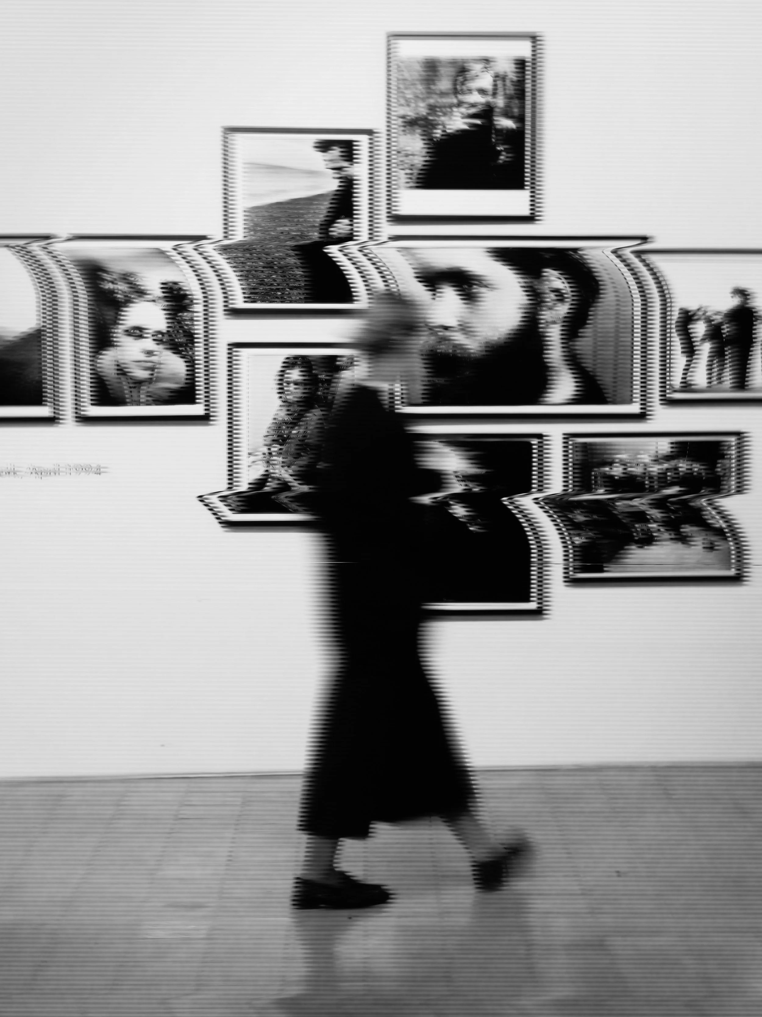Person walking in front of a wall with framed photographs, blurry and distorted in black and white.