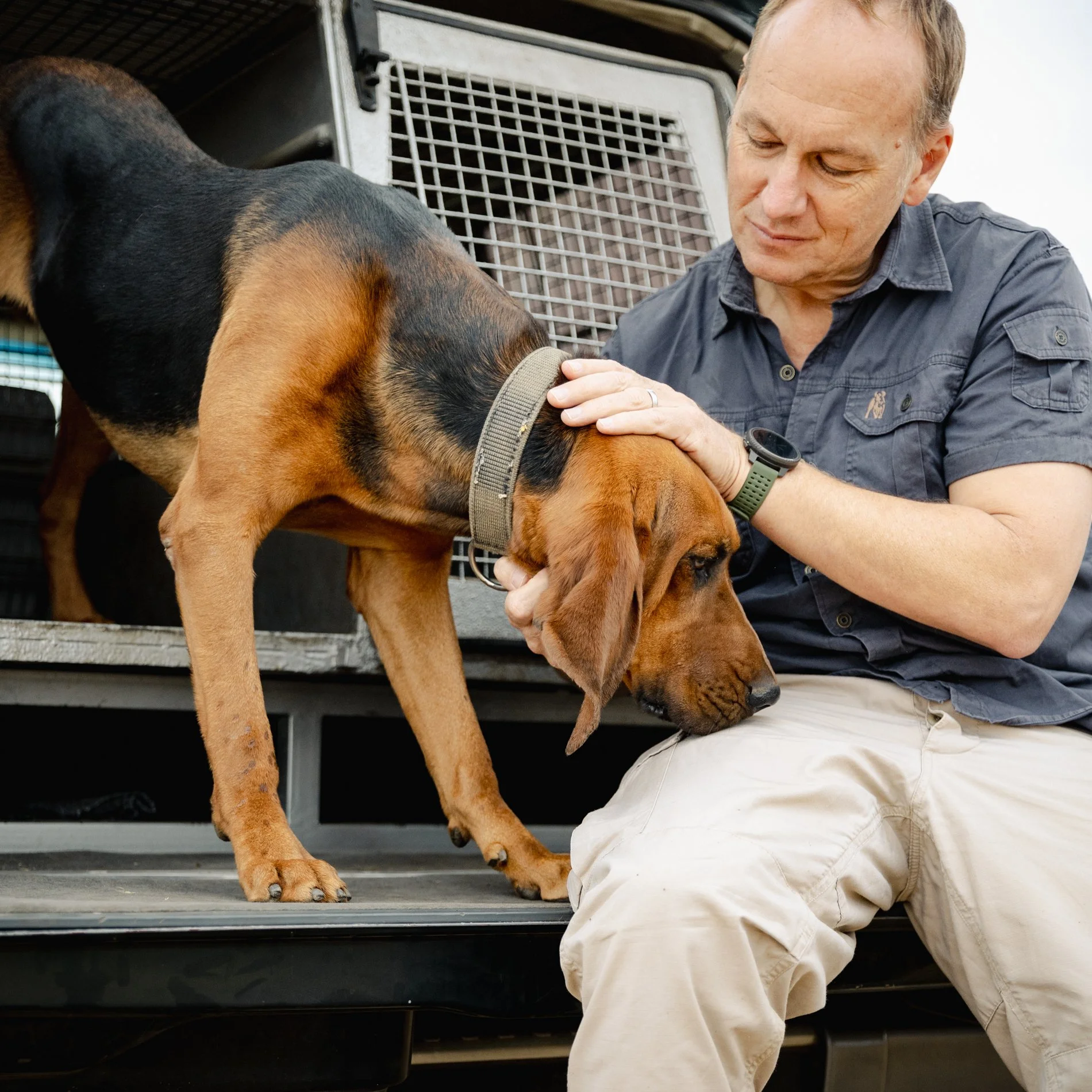 A man kneeling next to a large dog, possibly a bloodhound, in front of a trailer with a metal grate door. The man is gently touching the dog's face and has a focused expression.