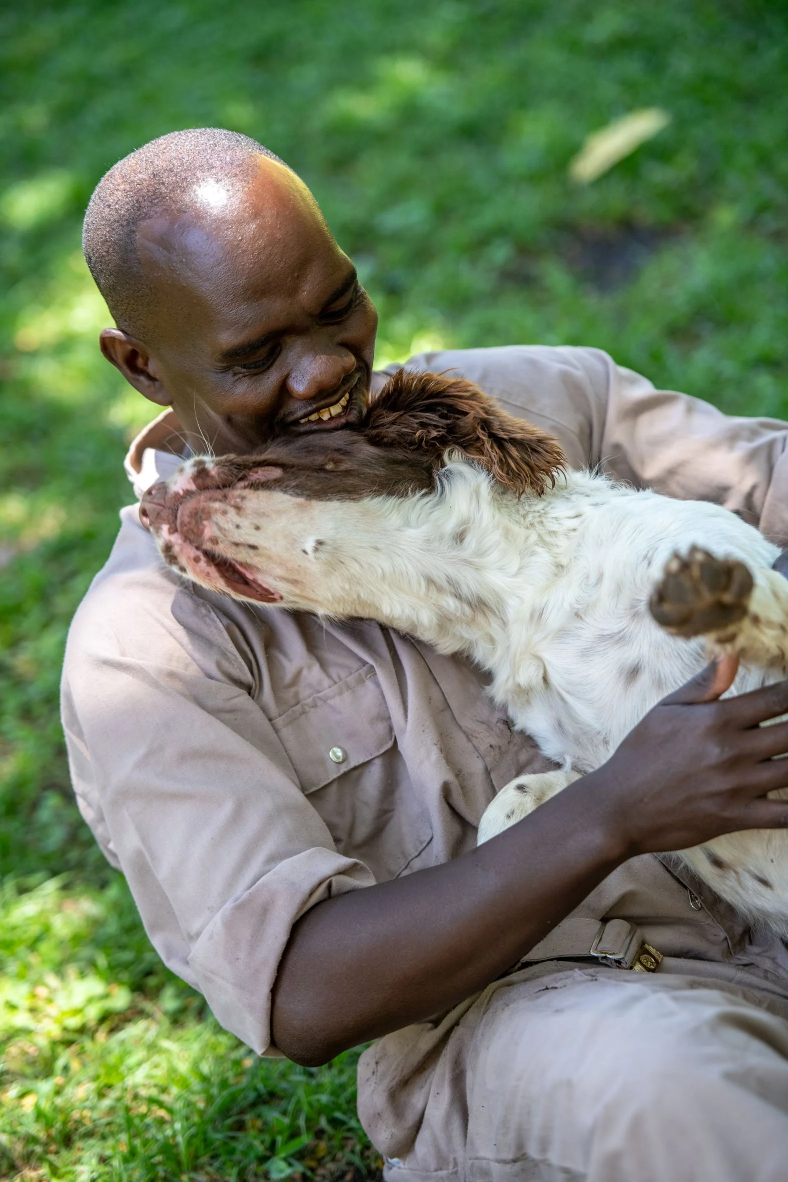 A man in a beige uniform sitting outdoors on green grass, hugging and smiling at a brown and white dog.