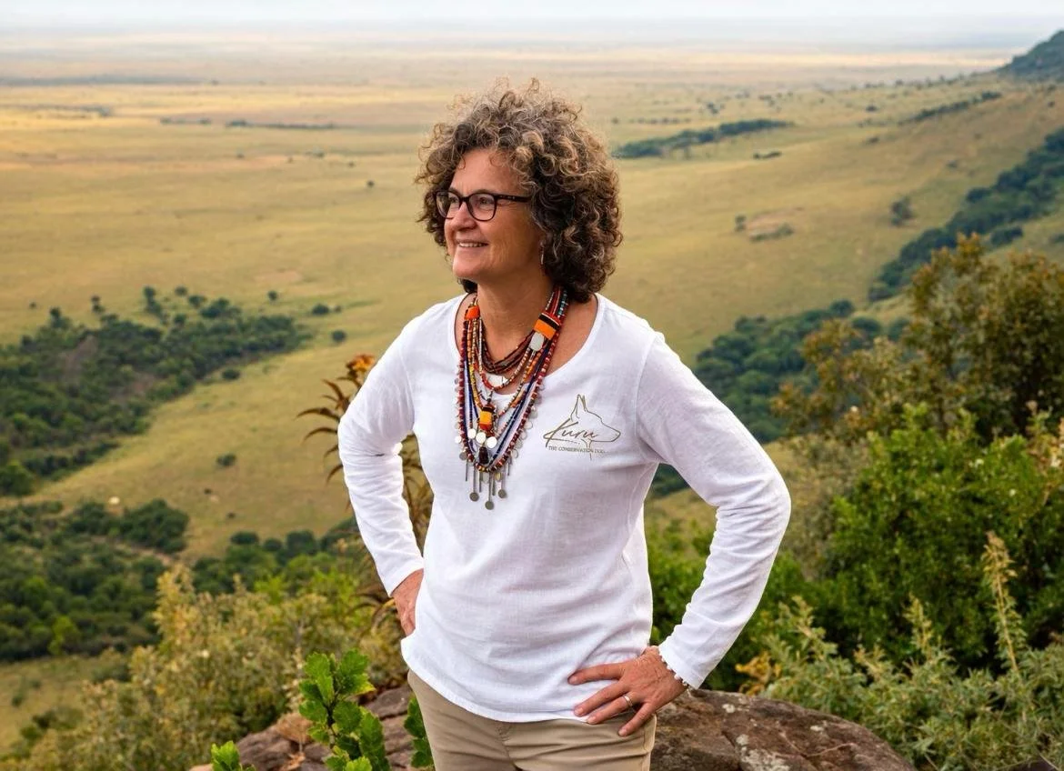 A woman with curly hair and glasses standing outdoors on a hillside, smiling and looking to the side, with a vast landscape of hills and greenery in the background.