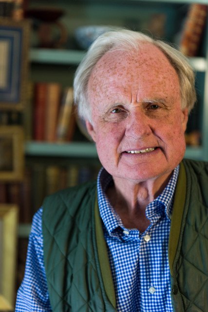 An elderly man with gray hair and a friendly smile, wearing a blue checkered shirt and a green vest, standing in a room with bookshelves and framed pictures in the background.