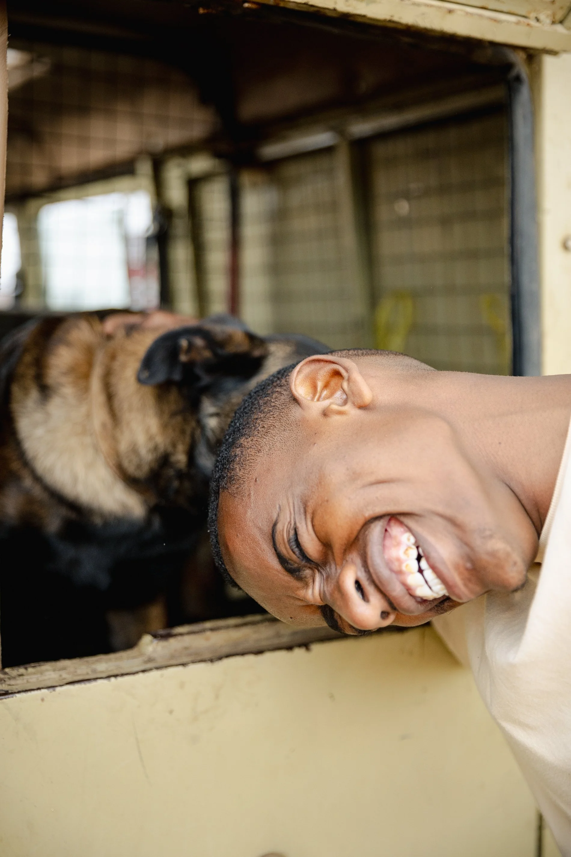 A man with short dark hair is smiling widely with his eyes closed, leaning his head against a dog. The dog is a German Shepherd with black and tan fur, resting its head on the man's shoulder. They are inside a room with a metal-wired cage or kennel in the background.