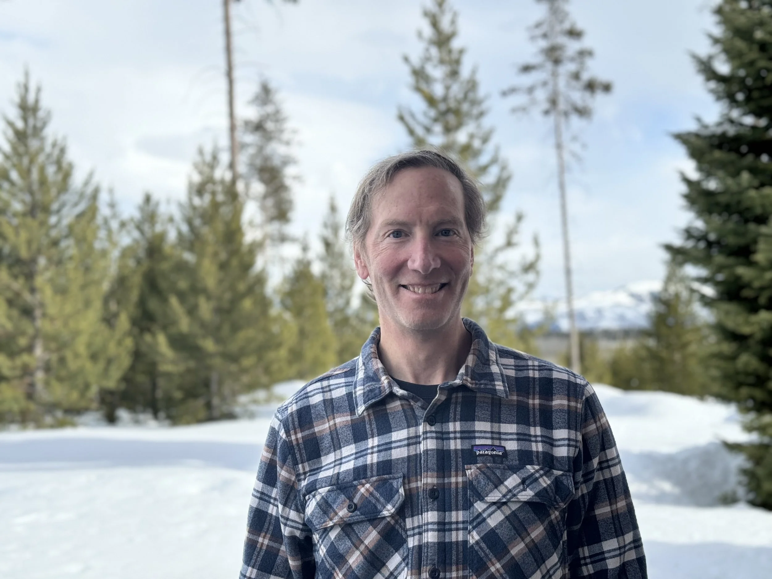 A smiling man wearing a plaid flannel shirt standing outdoors in a snowy landscape with trees in the background.