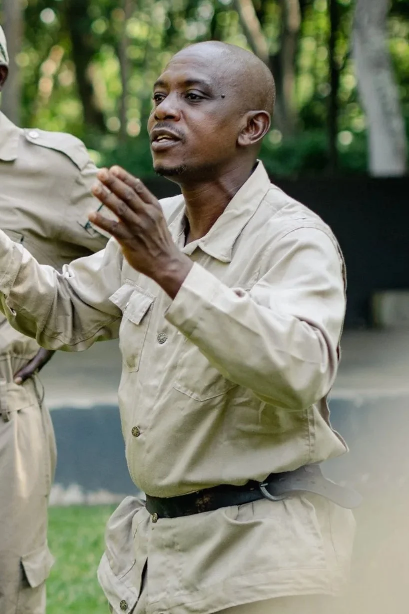A man in a beige uniform gesturing while talking outdoors with trees in the background.