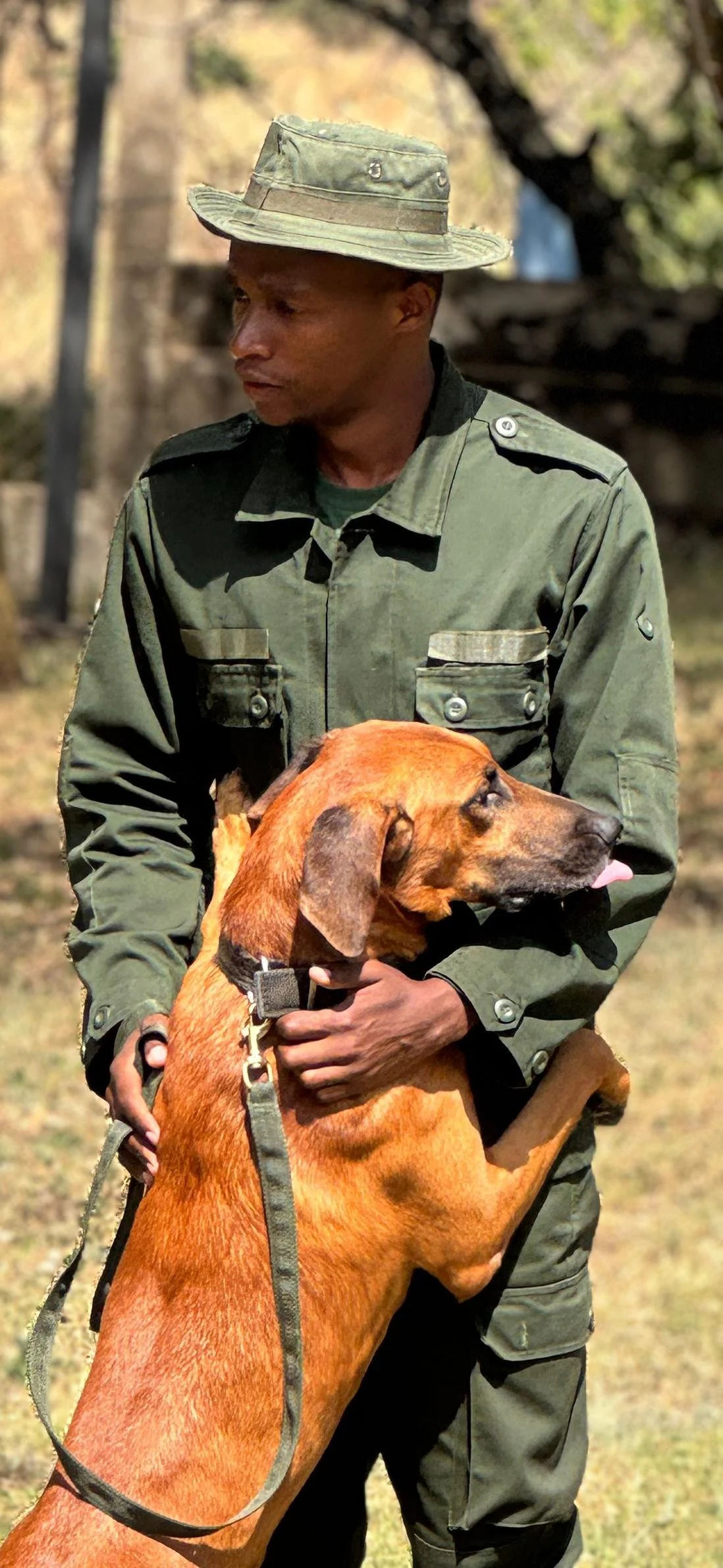 A man in military uniform holding a brown dog outdoors, with a blurred background of trees and grass.
