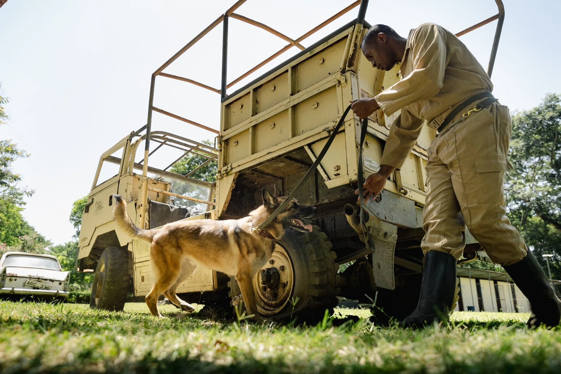 A man in uniform with a dog on a leash standing near a yellow military-style vehicle outdoors, with trees and a building in the background.