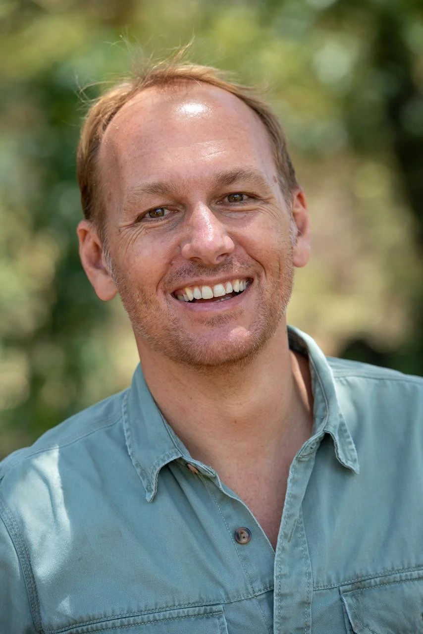 A smiling man with light brown hair and stubble, wearing a light green button-up shirt, outdoors with a blurred background of trees and greenery.