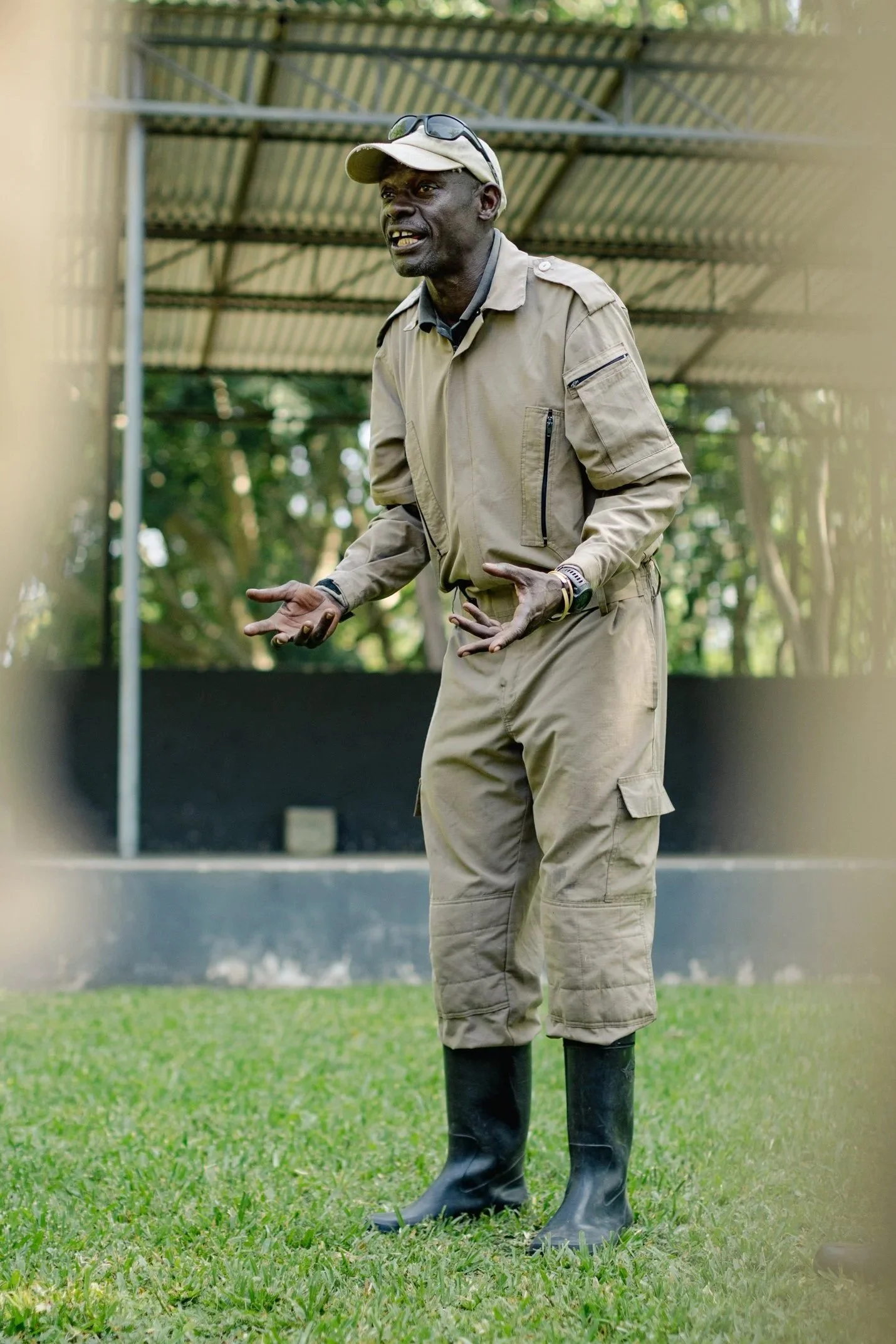 A man dressed in beige outdoor clothing, including a cap, jacket, and cargo pants, standing on a grassy field with trees in the background. He is wearing black rain boots and appears to be speaking or giving instructions.