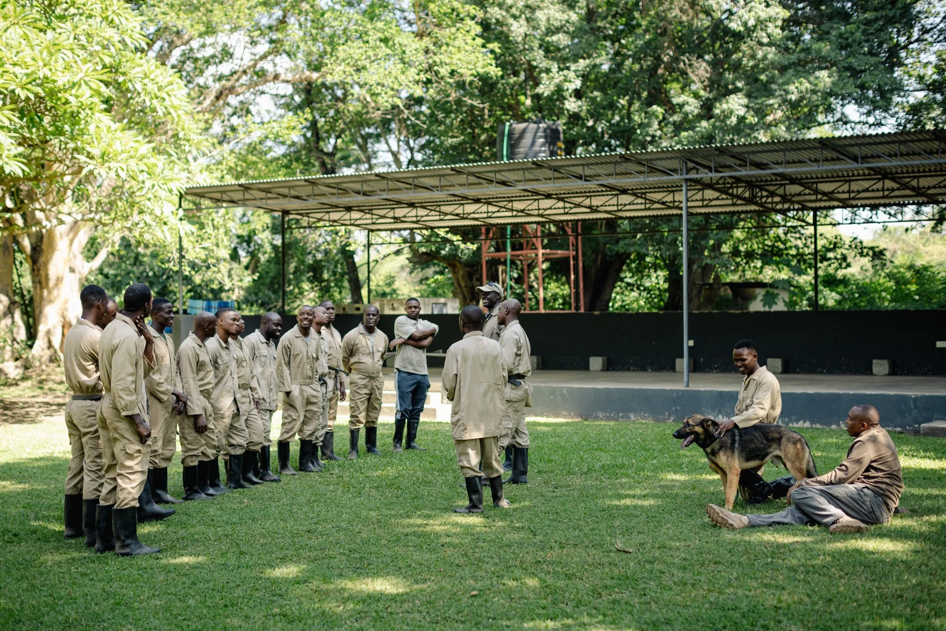 Group of people in uniform listening to an instructor outdoors under a metal shelter. Some are sitting or standing, with a dog present. Trees in the background and a shaded grassy area.