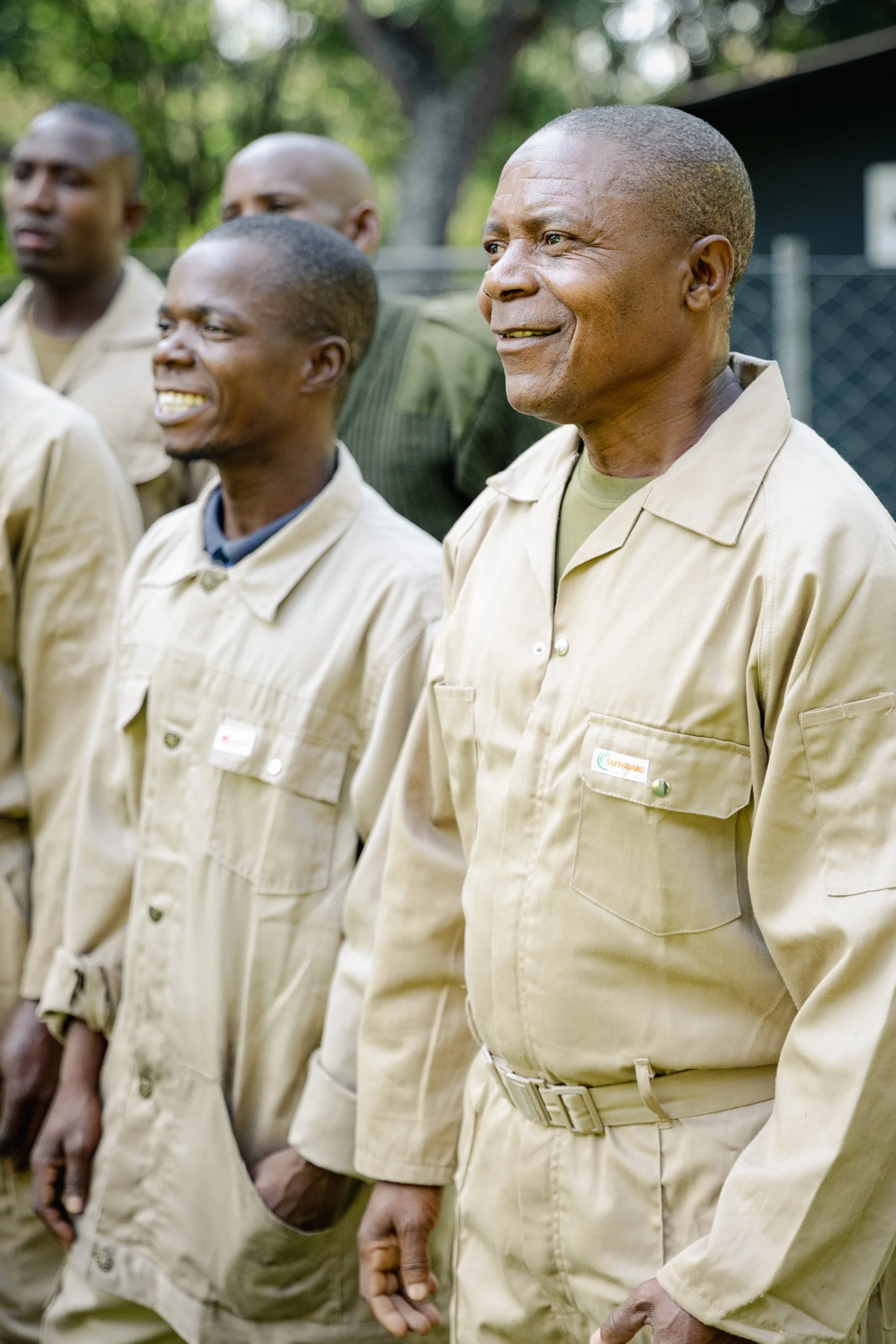 A group of men standing outdoors, dressed in beige prison jumpsuits with name tags, in a line with serious and content expressions.