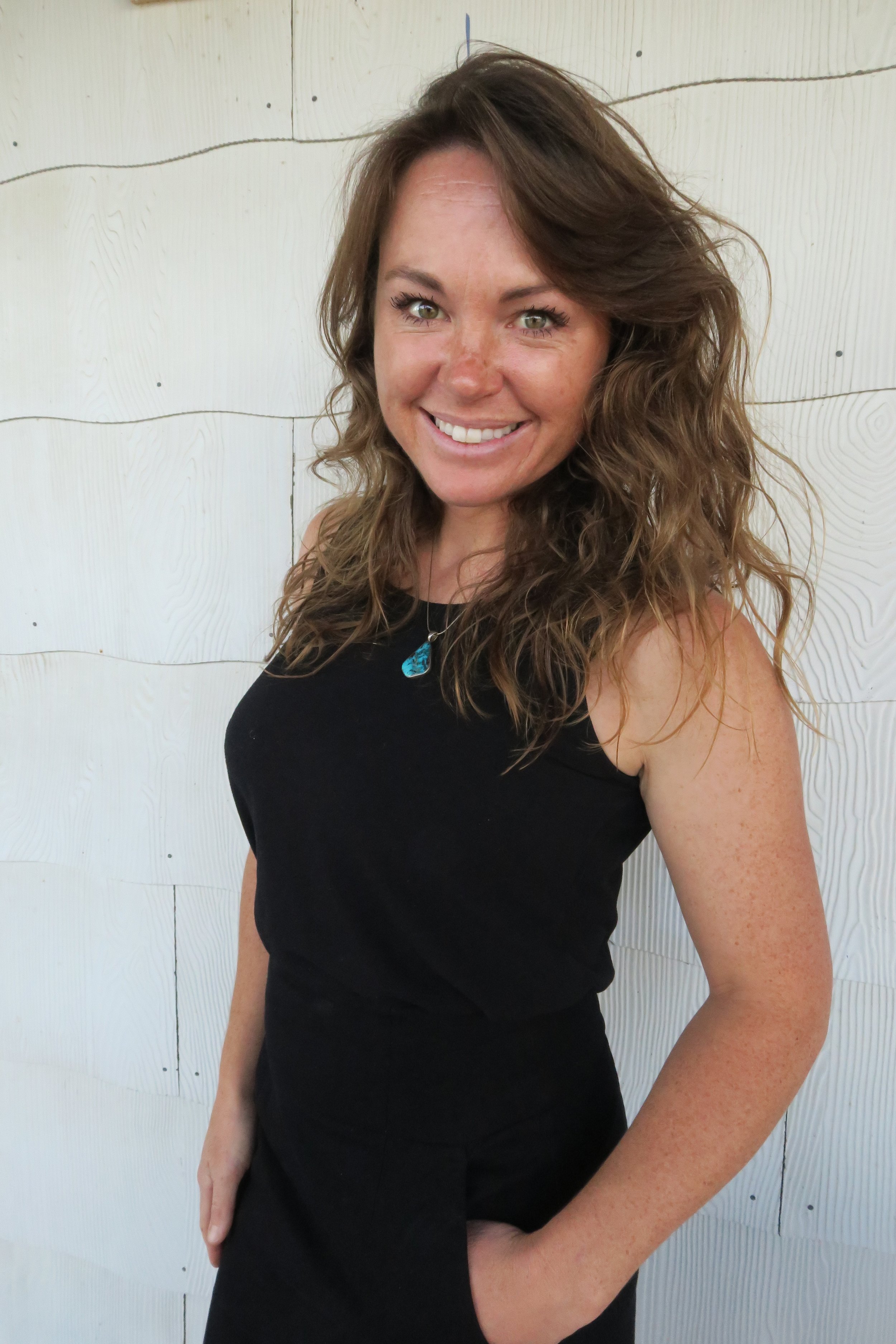 A woman with long curly brown hair wearing a black sleeveless top, standing against a light-colored wooden wall, smiling at the camera.