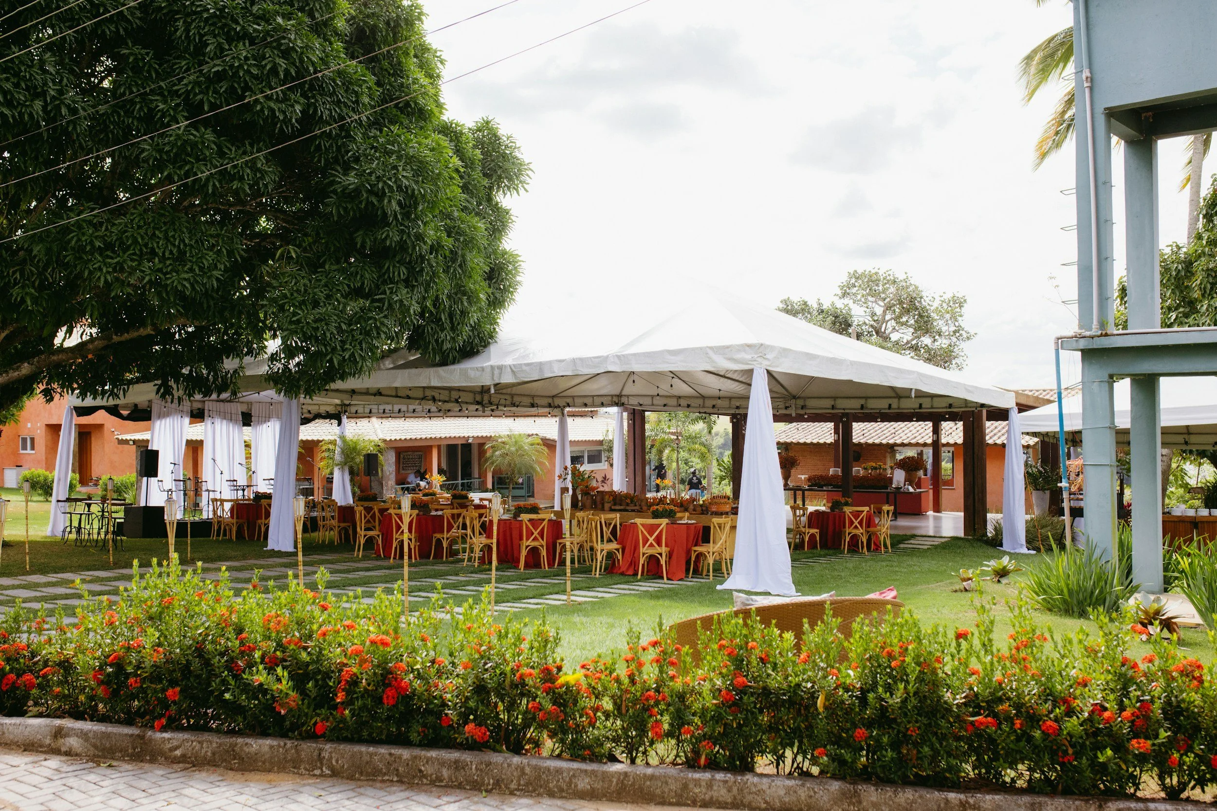 An outdoor event setup with a large white canopy tent, red tablecloths, and wooden chairs, surrounded by lush greenery and a flowerbed in the foreground.