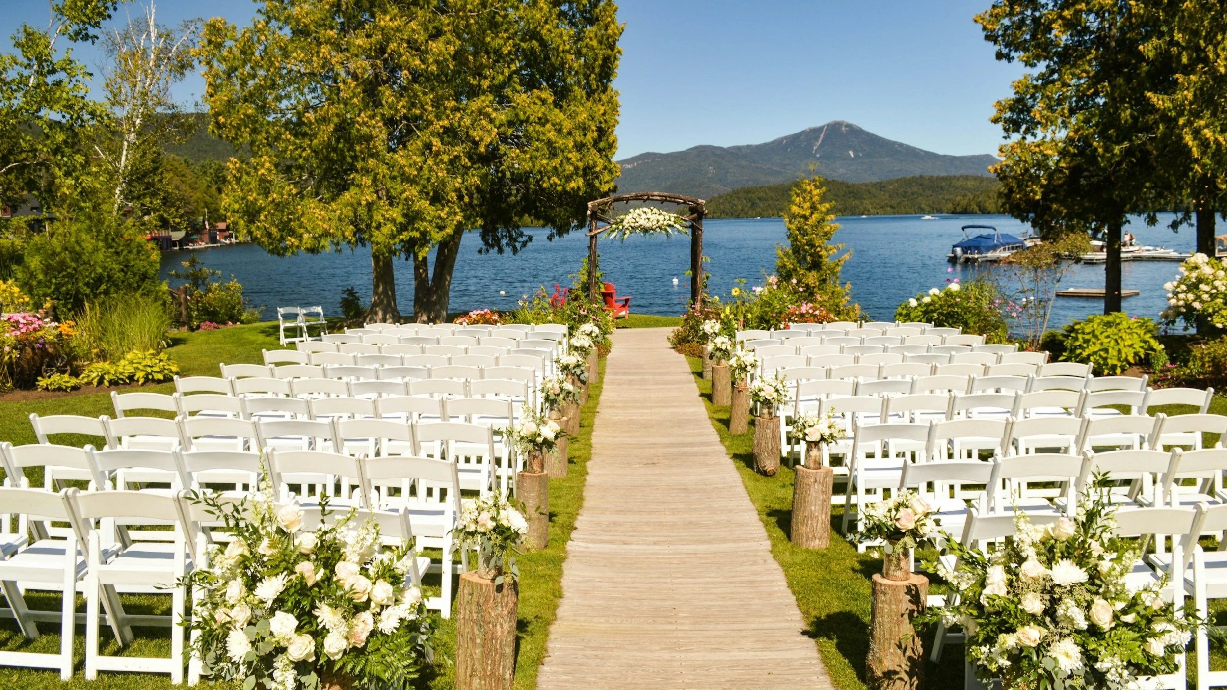 An outdoor wedding setup with white chairs decorated with flowers, a wooden aisle, and a view of a lake with boats and mountains in the background.